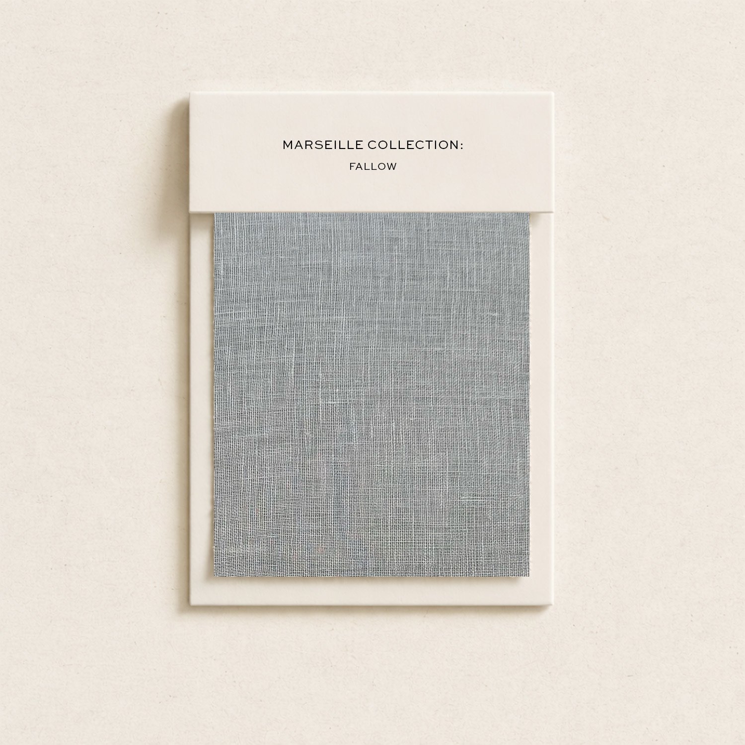 A sample of gray fabric with a white border displayed in a white frame and a white label at the top that reads 'Marseille Collection: Fallow' against a light beige wall.