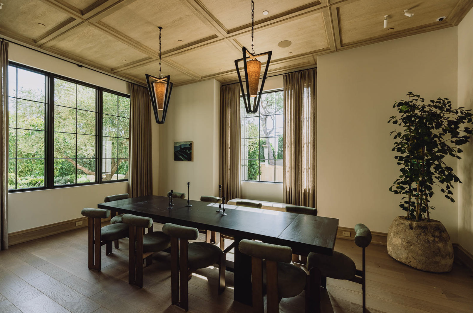 A dining room with a dark wooden table surrounded by eight upholstered chairs, large windows with brown curtains, and hanging pendant lights, with trees visible outside.