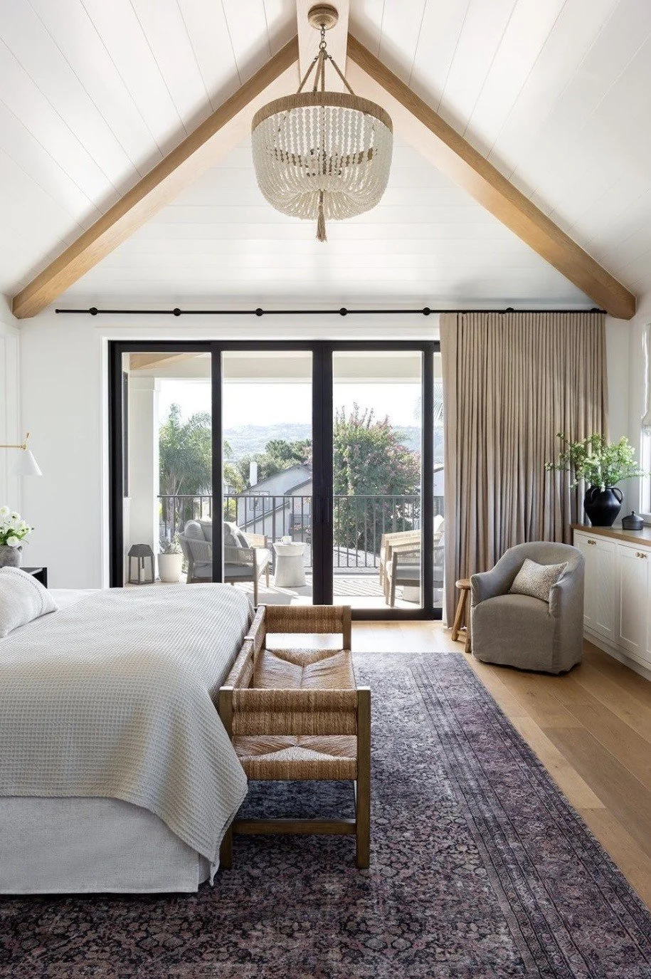 Bright bedroom with vaulted ceiling, white walls, and a large sliding glass door opening to a balcony with outdoor seating. The room features a white bed with a textured blanket, a wooden bench at the foot, a gray armchair with a pillow, a dresser wi
