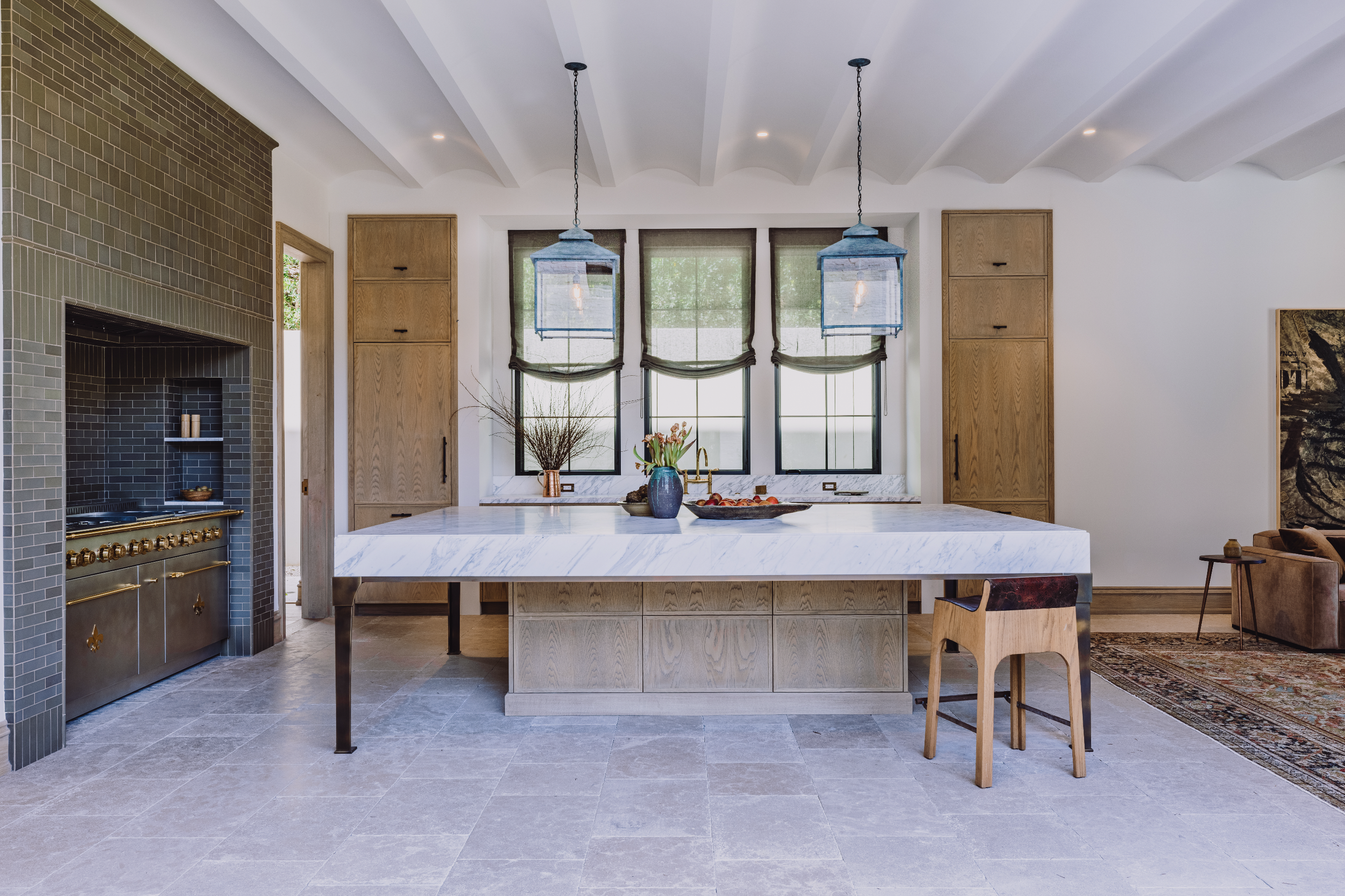 Modern kitchen with white marble island, wooden cabinets, large windows with green shades, and two blue pendant lights hanging from a white vaulted ceiling.
