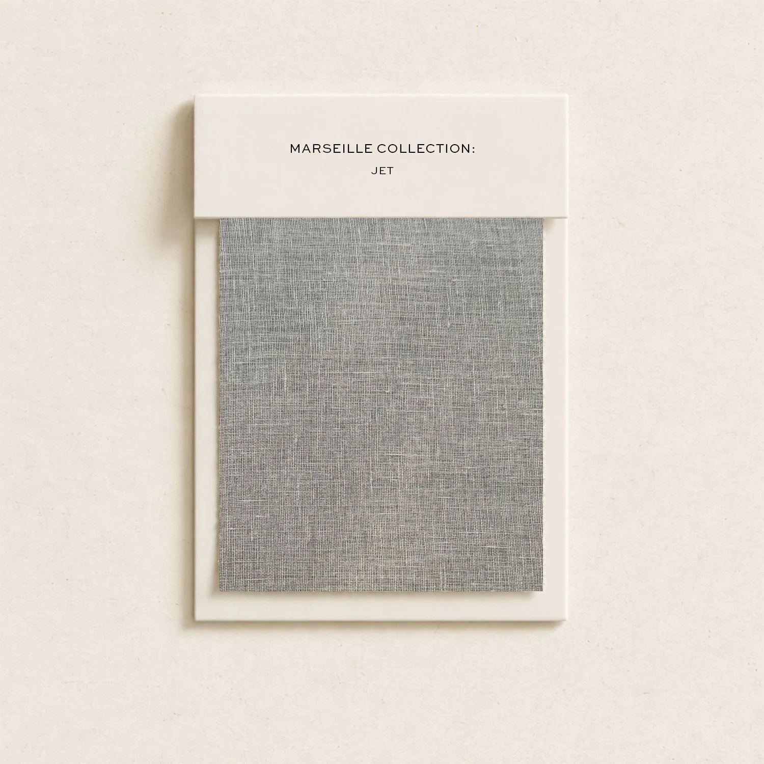 A fabric sample book titled 'Marseille Collection: Jet' with a gray textured fabric inside, mounted on a white backing against a light-colored wall.