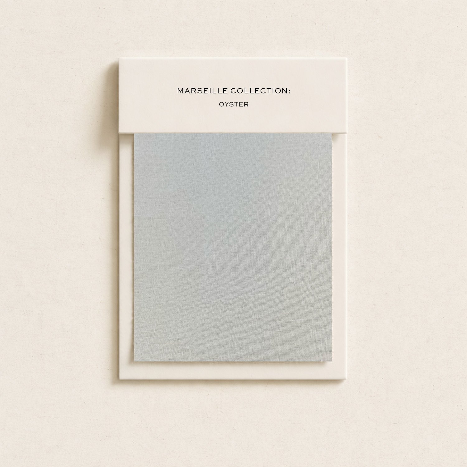 A white frame holding a sample of light gray fabric with a label at the top reading 'Marseille Collection: Oyster' on a beige wall.