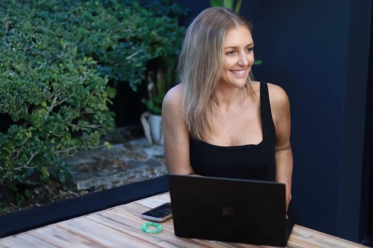 A woman with blonde hair sitting outdoors at a table with a laptop and a smartphone, smiling.