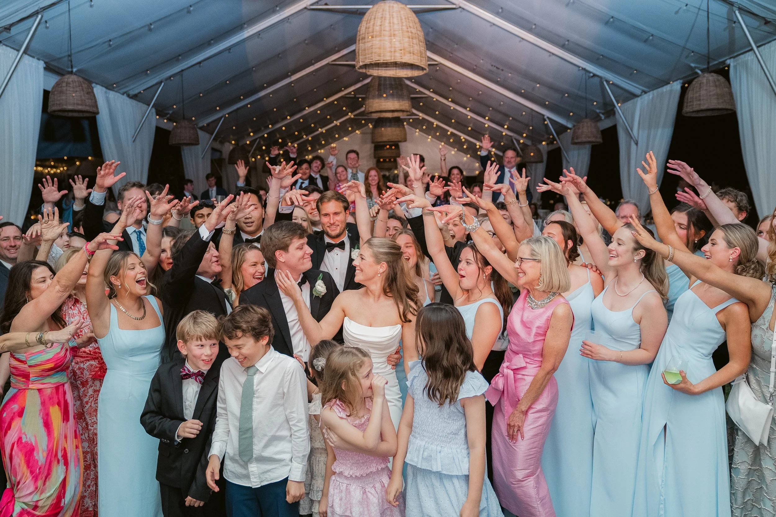 A large group of people at a celebration, many dressed in formal and colorful attire, some in suits and ties, celebrating in a decorated venue with hanging lights and curtains.