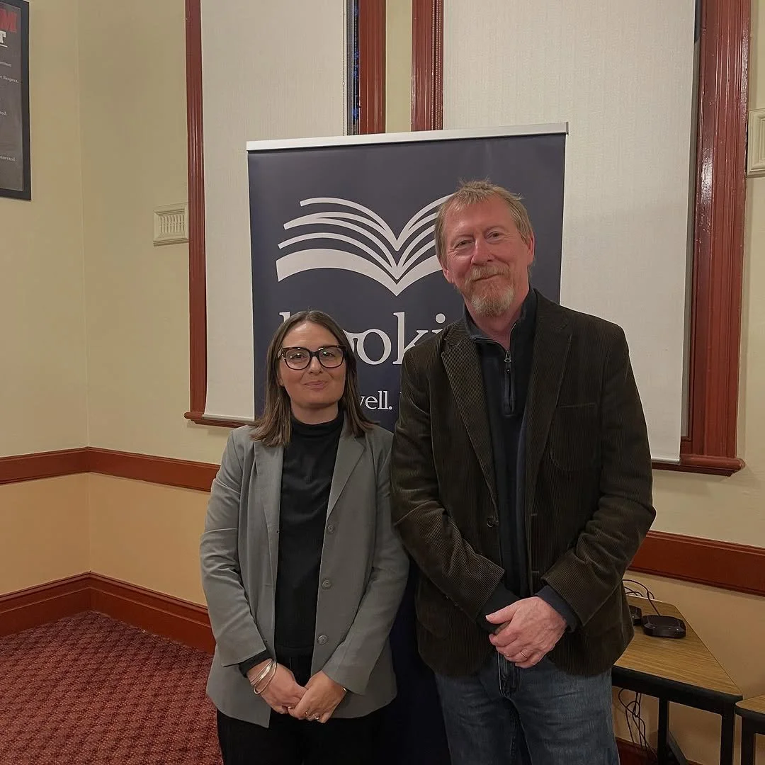 A woman and a man standing indoors in front of a banner with an open book logo. The woman is wearing glasses and a gray blazer, while the man has a beard and is wearing a dark jacket. Both are smiling at the camera.