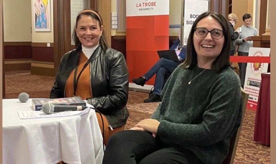 Two women sitting at a table with microphones in front of them, smiling at the camera. They are in a conference or event setting with a banner for La Trobe University behind them.