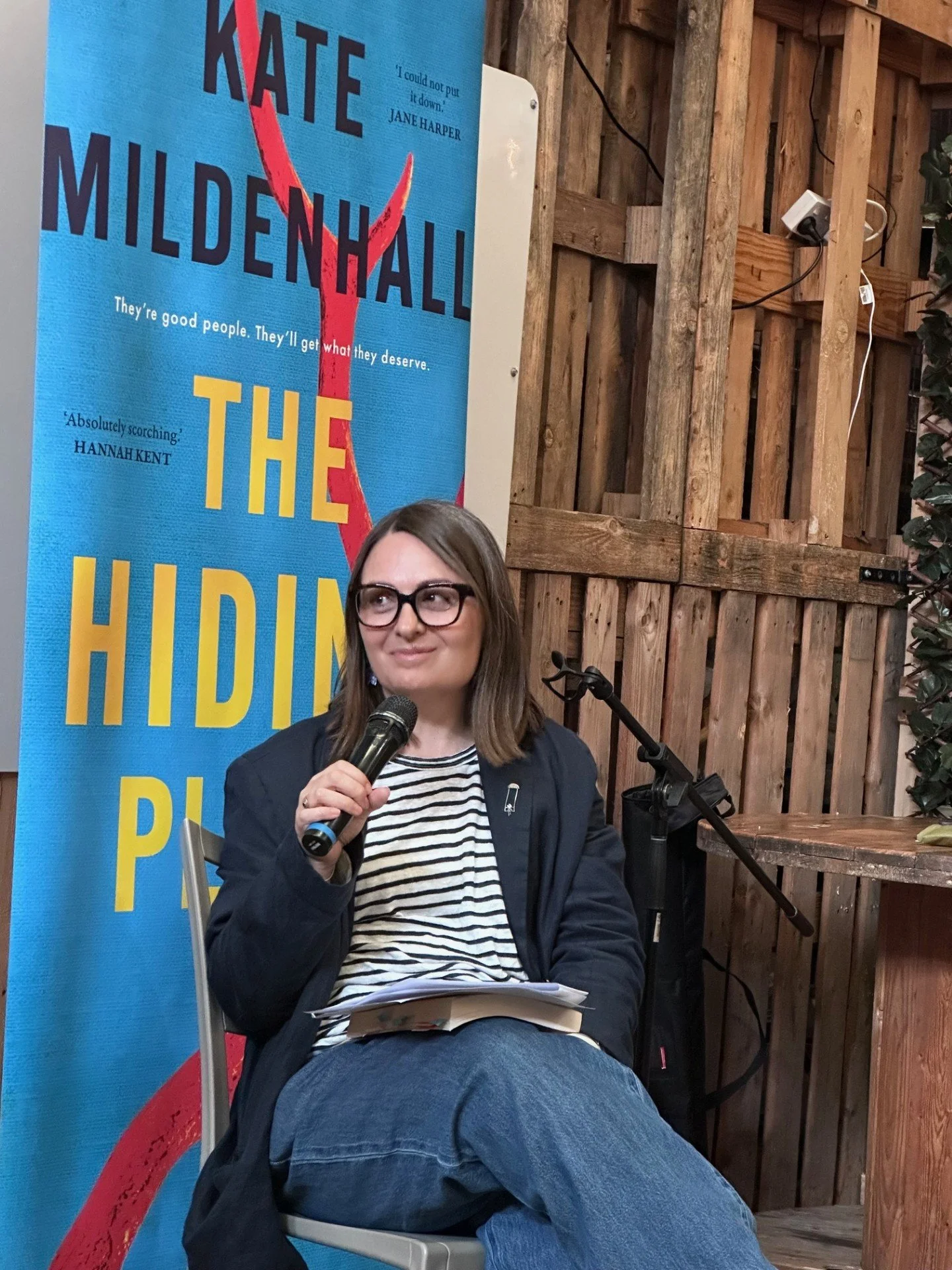 A woman with glasses and shoulder-length hair sitting on a chair, holding a microphone, with a slogan poster for 'The Hiding Place' in the background.