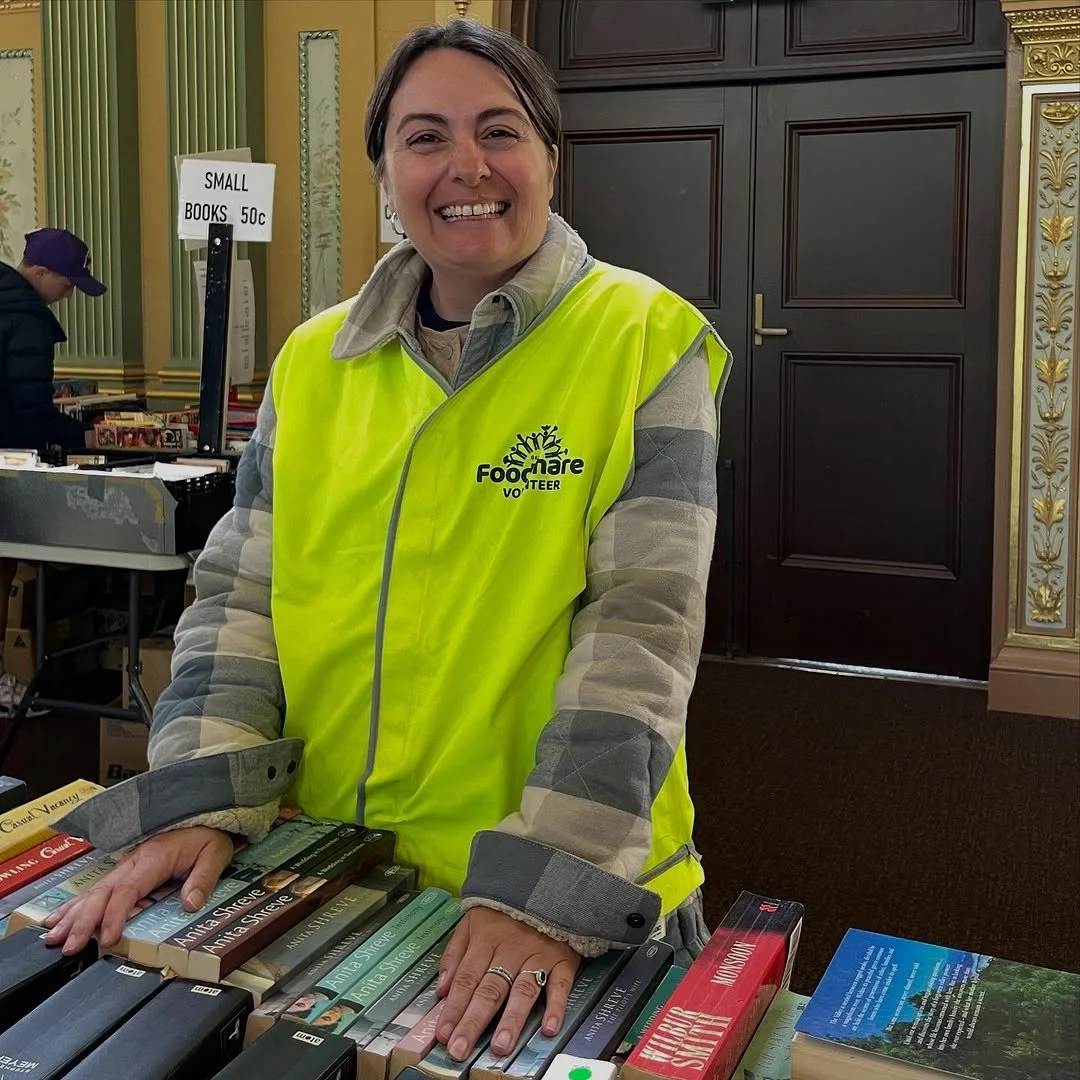 A smiling woman wearing a yellow volunteer vest stands behind a table with books at a book sale event.
