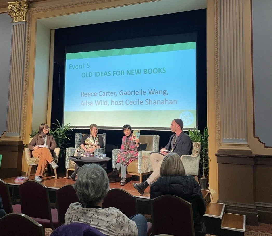 Panel discussion at a literary event with four women and one man on stage, seated in armchairs, with a large screen behind them displaying the event title and speakers, in a theater with ornate decor.