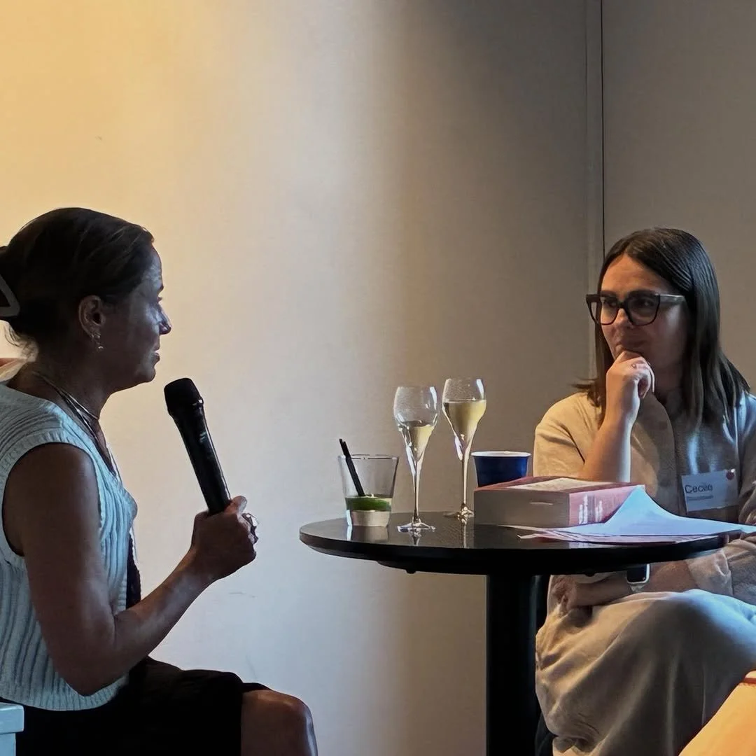 Two women seated at a round table engaged in conversation. The woman on the left holds a microphone, and the woman on the right, wearing glasses, listens thoughtfully. The table has three glasses of champagne, a glass with a lime wedge, some papers, a book, a blue cup, and a pink notepad. The setting appears to be a professional or conference environment.