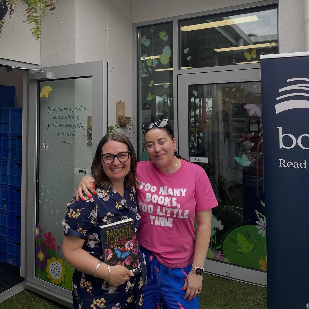 Two women smiling and standing outside a building with glass windows and decorated with flower and butterfly stickers. One woman is holding a book titled 'Rachel Johns'.