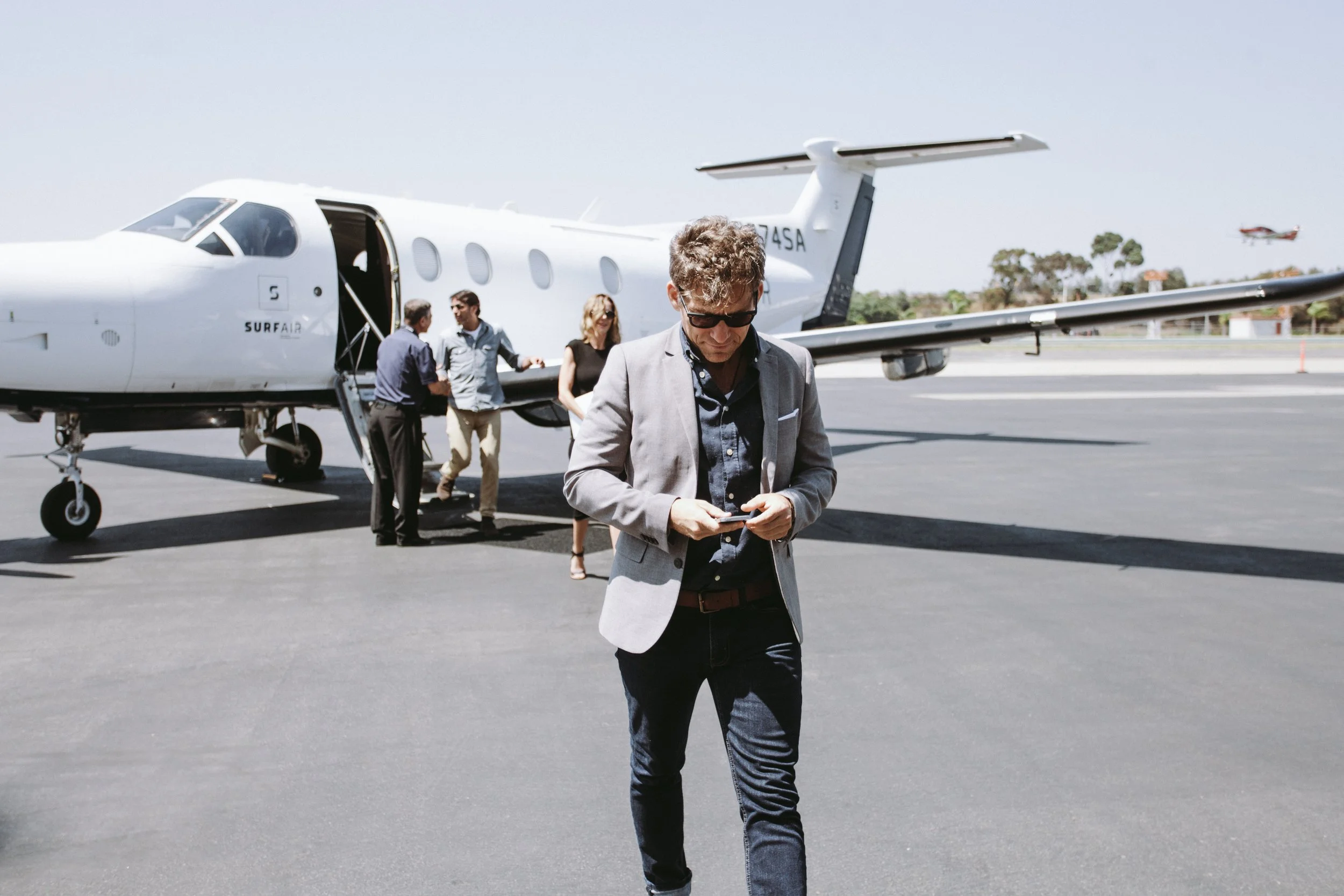 A man in a gray blazer and sunglasses looking at his phone on an airport tarmac, with a small private jet and other passengers boarding in the background.