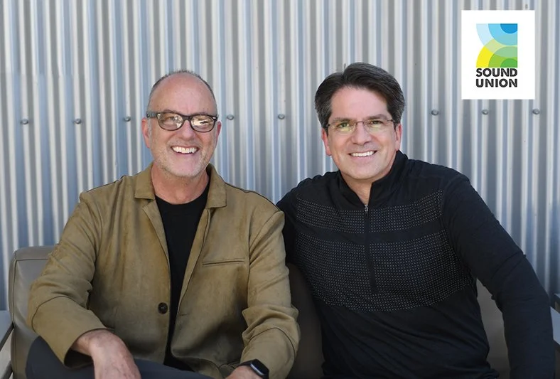 Two men sitting outdoors with a metal wall background, smiling at the camera, one wearing glasses and a tan jacket, the other in a black sweater. Sound Union logo in the top right corner.