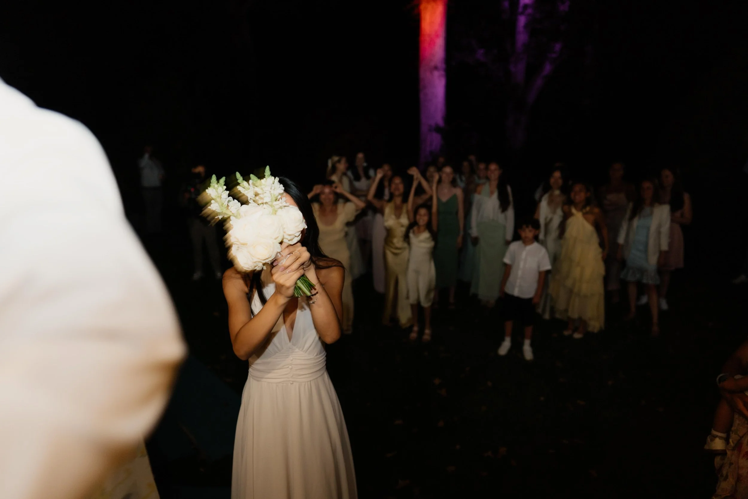 Frau trägt einen weißen Brautkleid und hält einen Blumenstrauß vor ihr Gesicht bei einer Brautfeier im Freien bei Nacht, im Hintergrund stehen Gäste und Bäume mit bunter Beleuchtung.