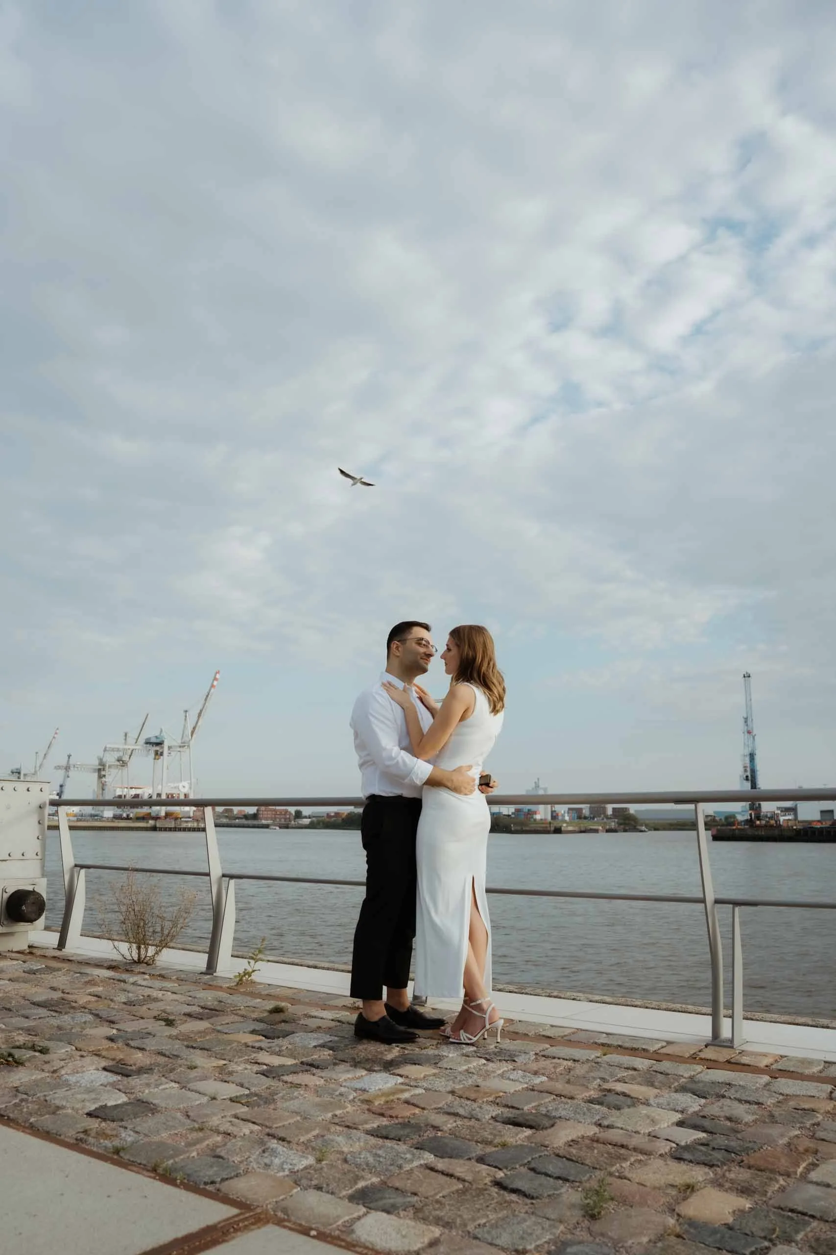Ein Paar in formeller Kleidung steht sich an einer Uferpromenade gegenüber, im Hintergrund Wasser, Container, Kräne und eine Stadt bei bewölktem Himmel, ein fliegender Vogel am Himmel.