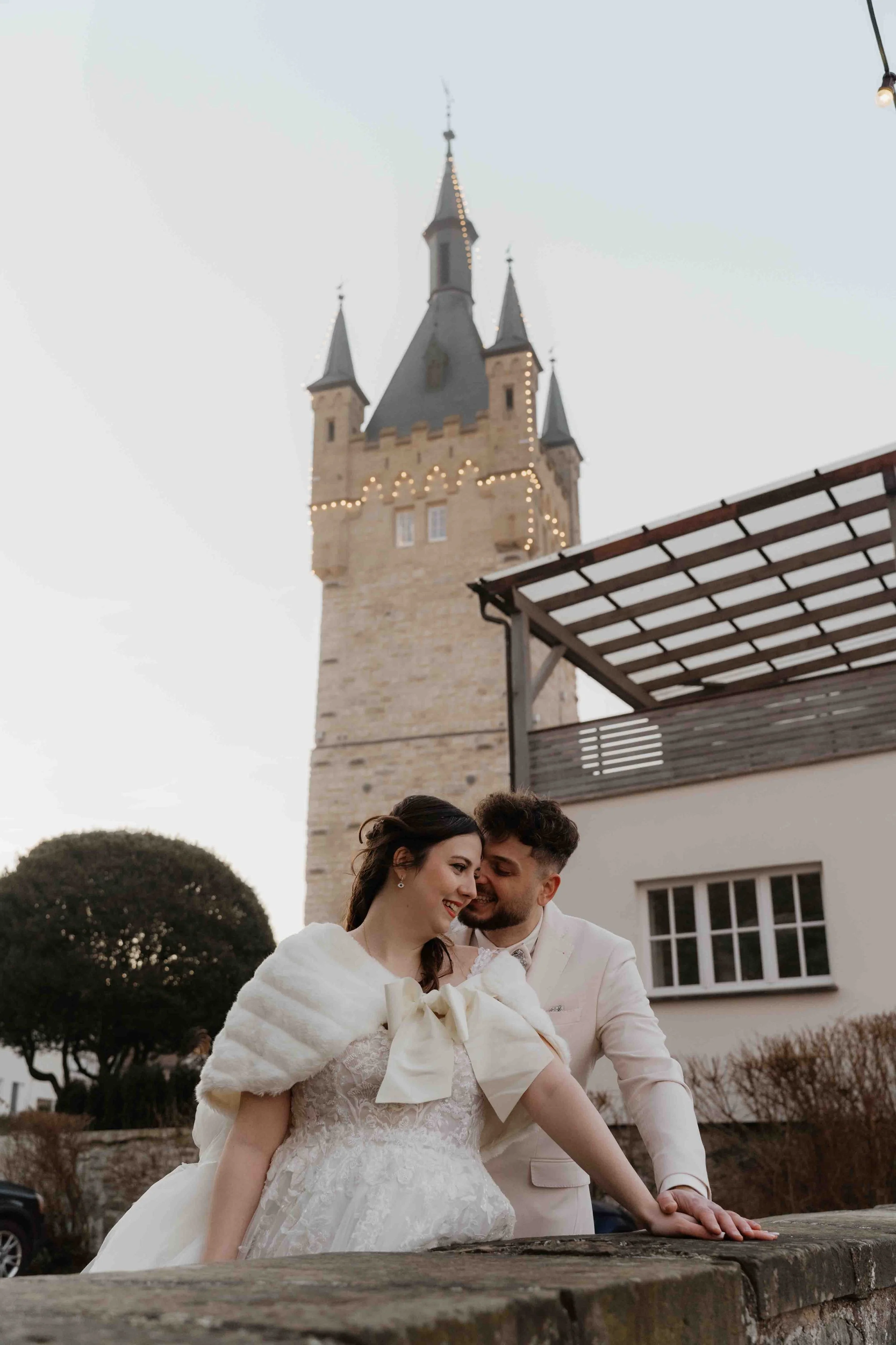 Ein frisch vermähltes Paar, die Braut trägt ein weißes Hochzeitskleid und einen weißen Umhang, der Bräutigam in einem hellen Anzug. Sie sind glücklich und lächeln sich an, während sie sich an einem Tag im Freien vor einer alten Burg oder einem Schlos