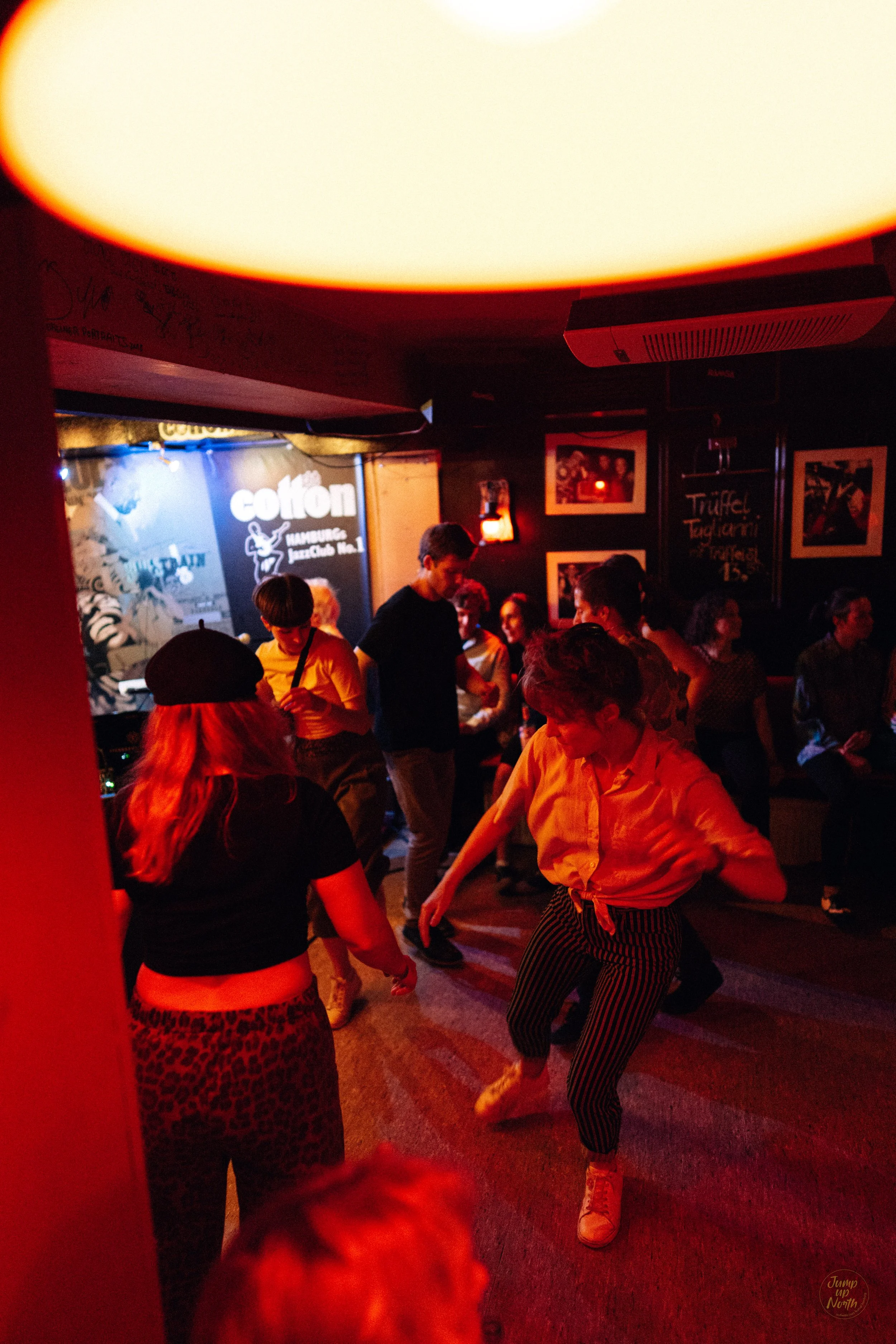 Menschen tanzen in einer Bar mit dunklen Wänden, Bilder an der Wand und gedämpftem Licht, das eine lebhafte Atmosphäre schafft.