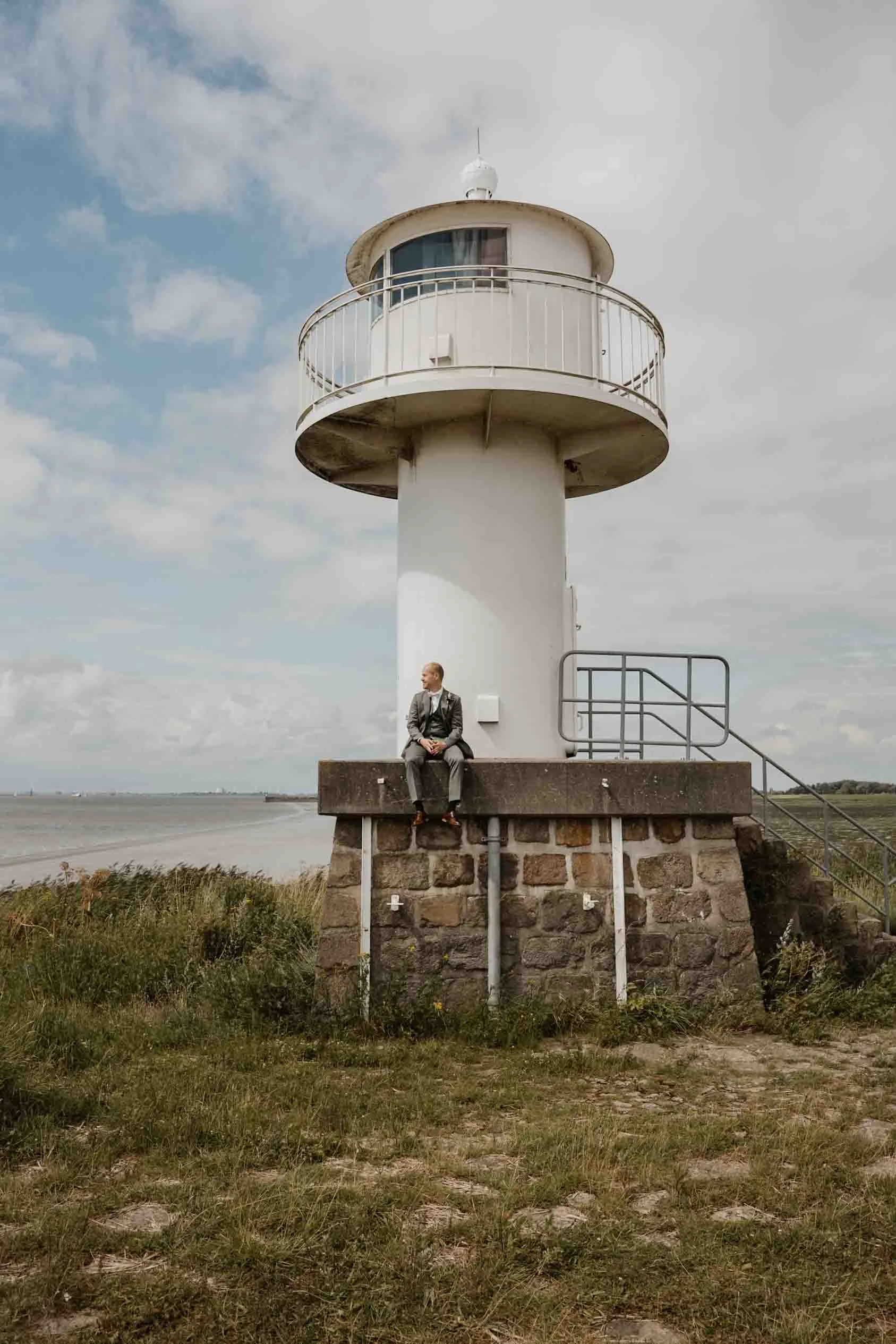 Eine weiße Leuchtturmszelle auf einem Steinfundament, mit einem Mann in einem Anzug, der davor sitzt, im Freien bei bewölktem Himmel und Meer im Hintergrund.