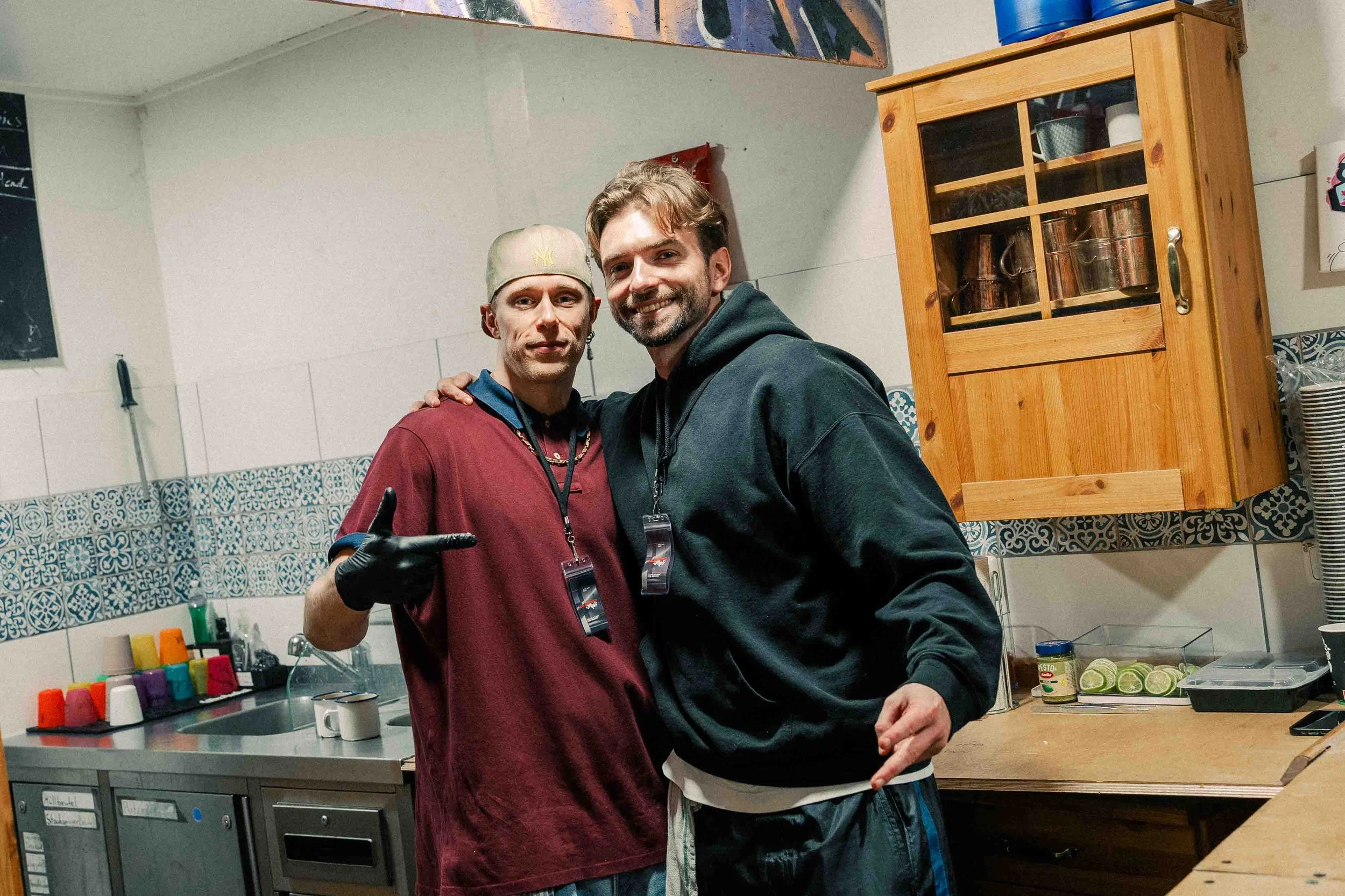 Zwei Männer stehen in einer Küche, beide lächeln und posieren für die Kamera. Der Mann auf der linken trägt eine beige Mütze, einen roten Pullover und schwarze Handschuhe. Der Mann auf der rechten trägt einen schwarzen Hoodie.