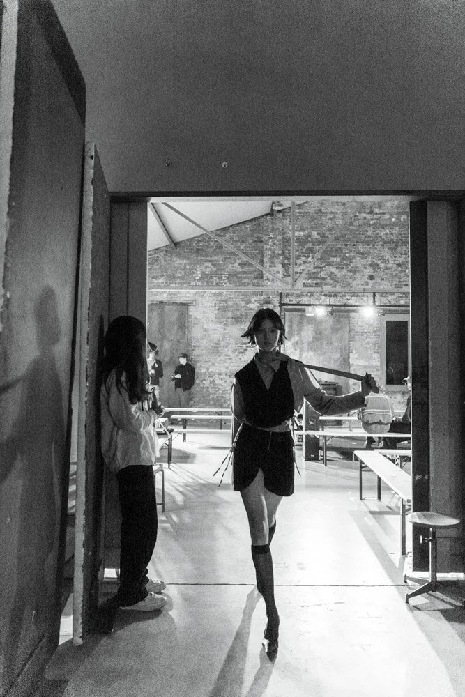Schwarz-weiß-Foto einer jungen Frau, die auf Stilettos in einem Kunst- oder Ausstellungsraum tanzt, während im Hintergrund zwei andere Personen stehen.