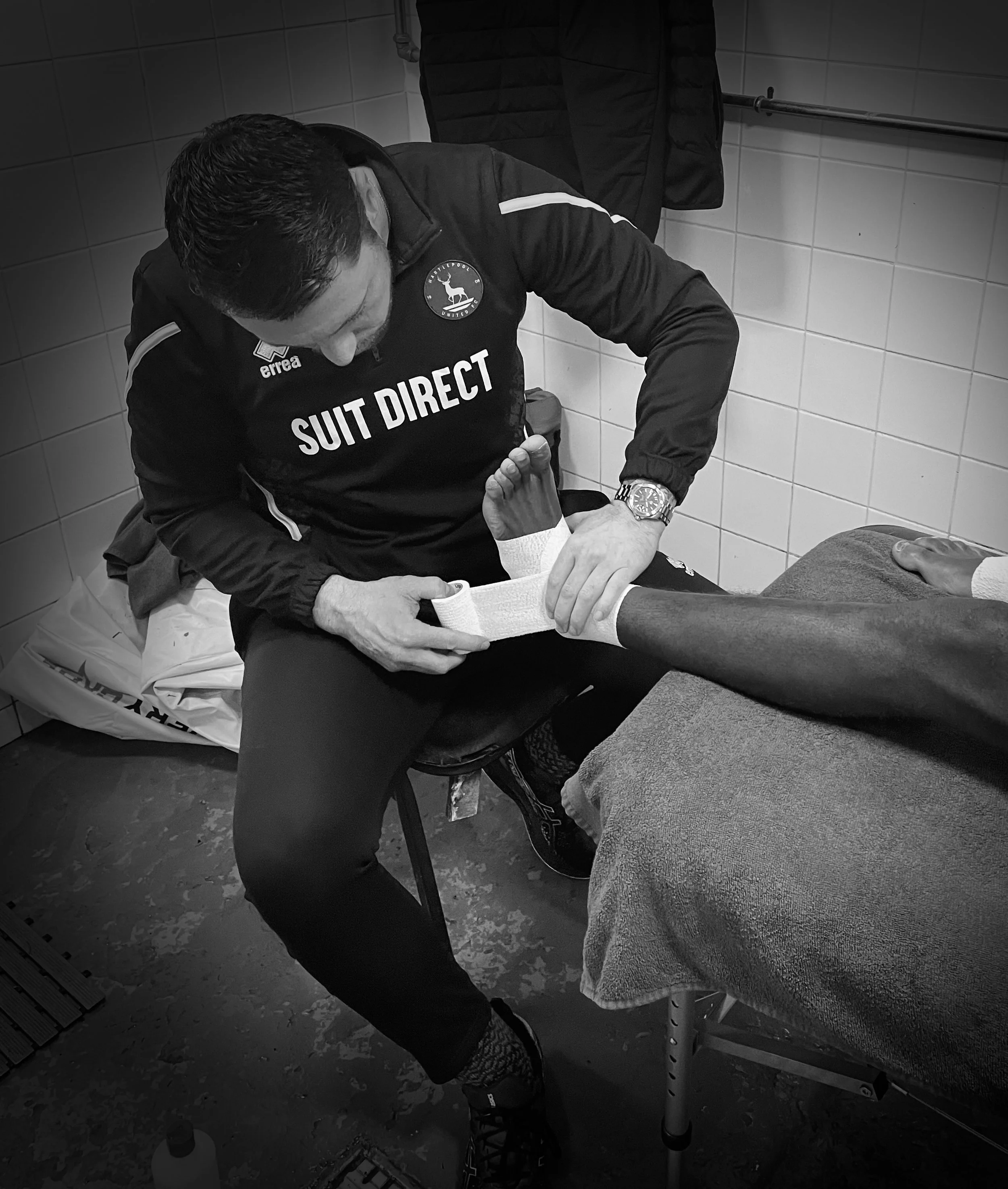 A man sitting on a stool in a tiled room, receiving medical attention to his foot from another man in a sports jacket with team logos and sponsor names.