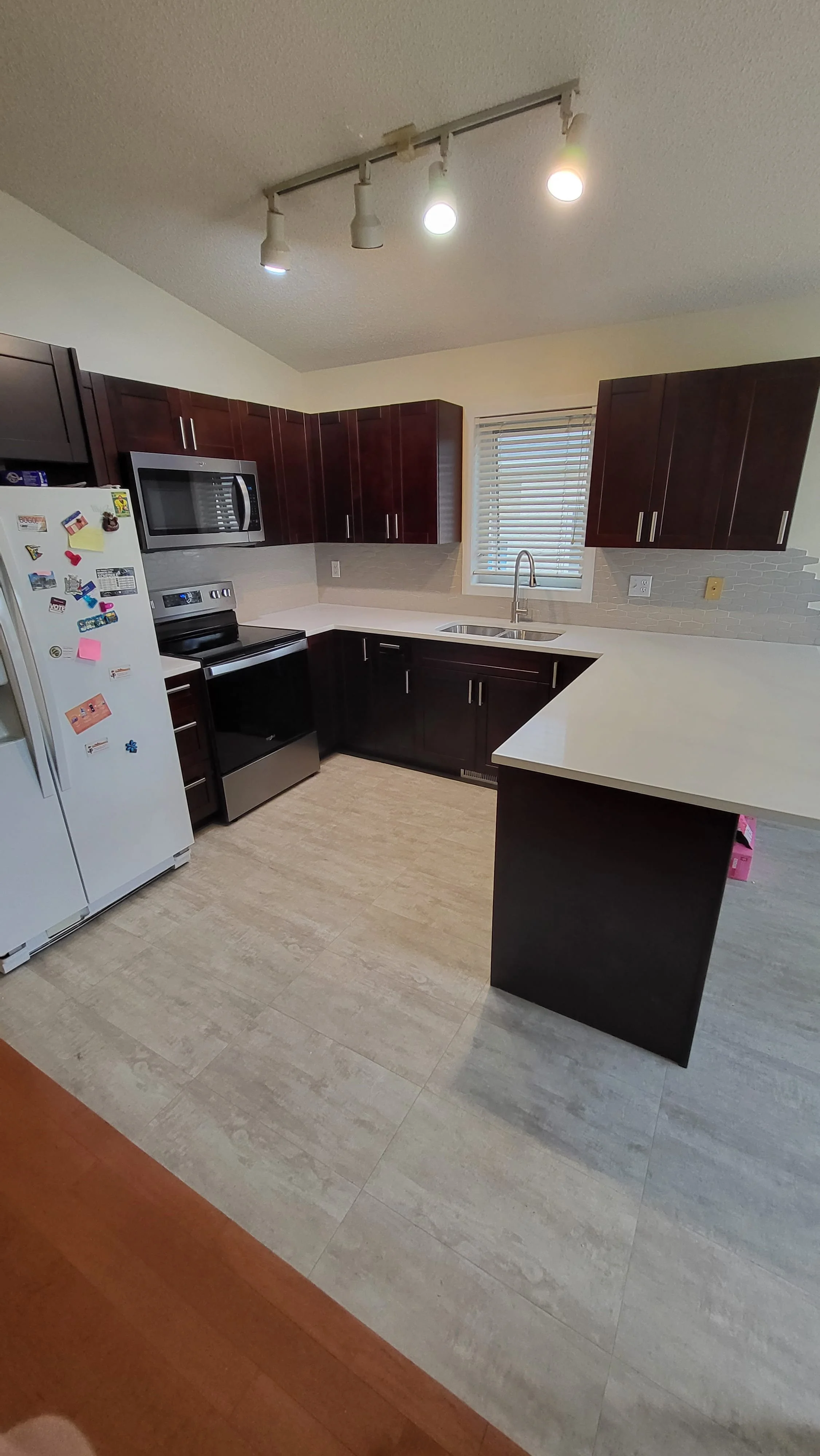 Kitchen with white countertops, dark wood cabinets, stainless steel appliances, a white refrigerator with magnets, a microwave, an oven, a sink under a window, and track lighting on the ceiling. kitchen contractor, cabinet installer, appliance instal