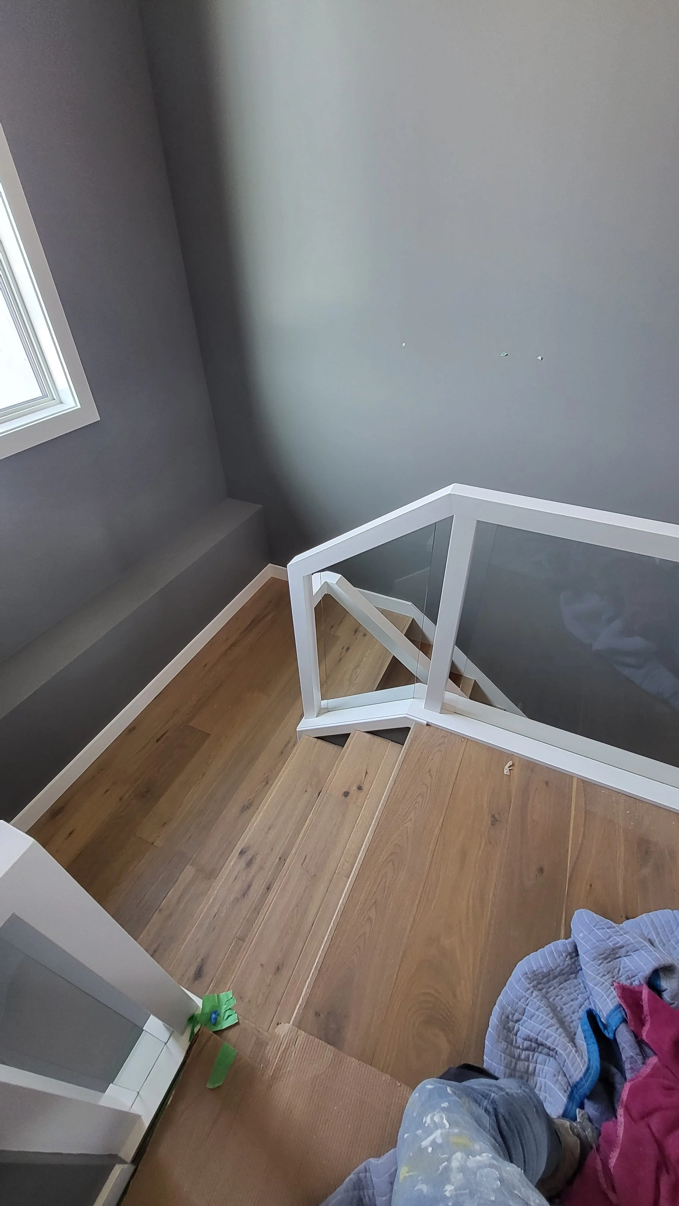Interior view of a staircase with wooden steps and a glass railing. The walls are painted gray, and there is a window on the left side. Some clothes and painting supplies are visible at the bottom right corner.  Basement Renovation, hardwood floors, 