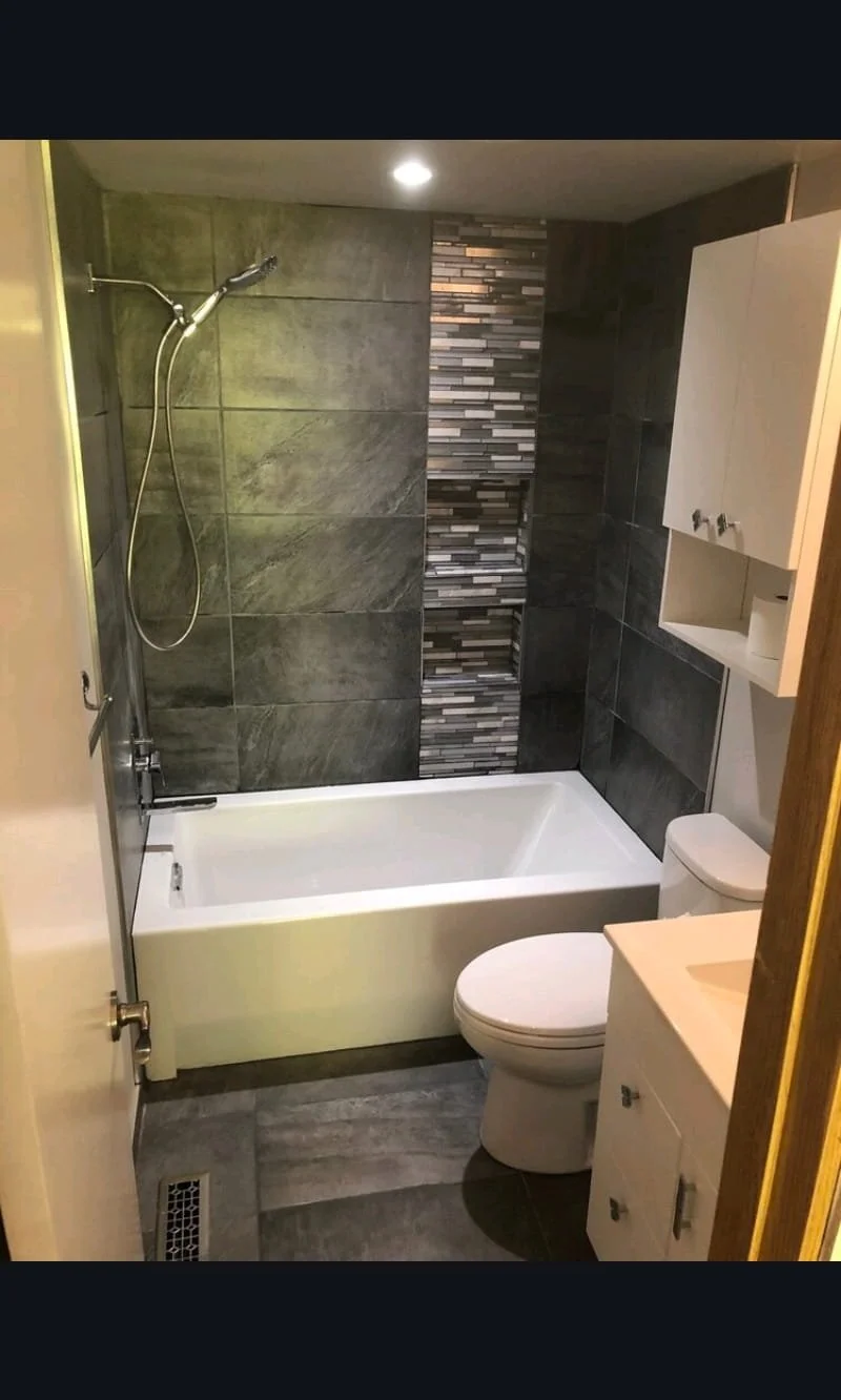 A small modern bathroom with a bathtub, a toilet, a wall-mounted cabinet, and a tiled wall with black and gray stone patterns. Bathroom contractor, tile installer, curb-less shower, glass shower doors