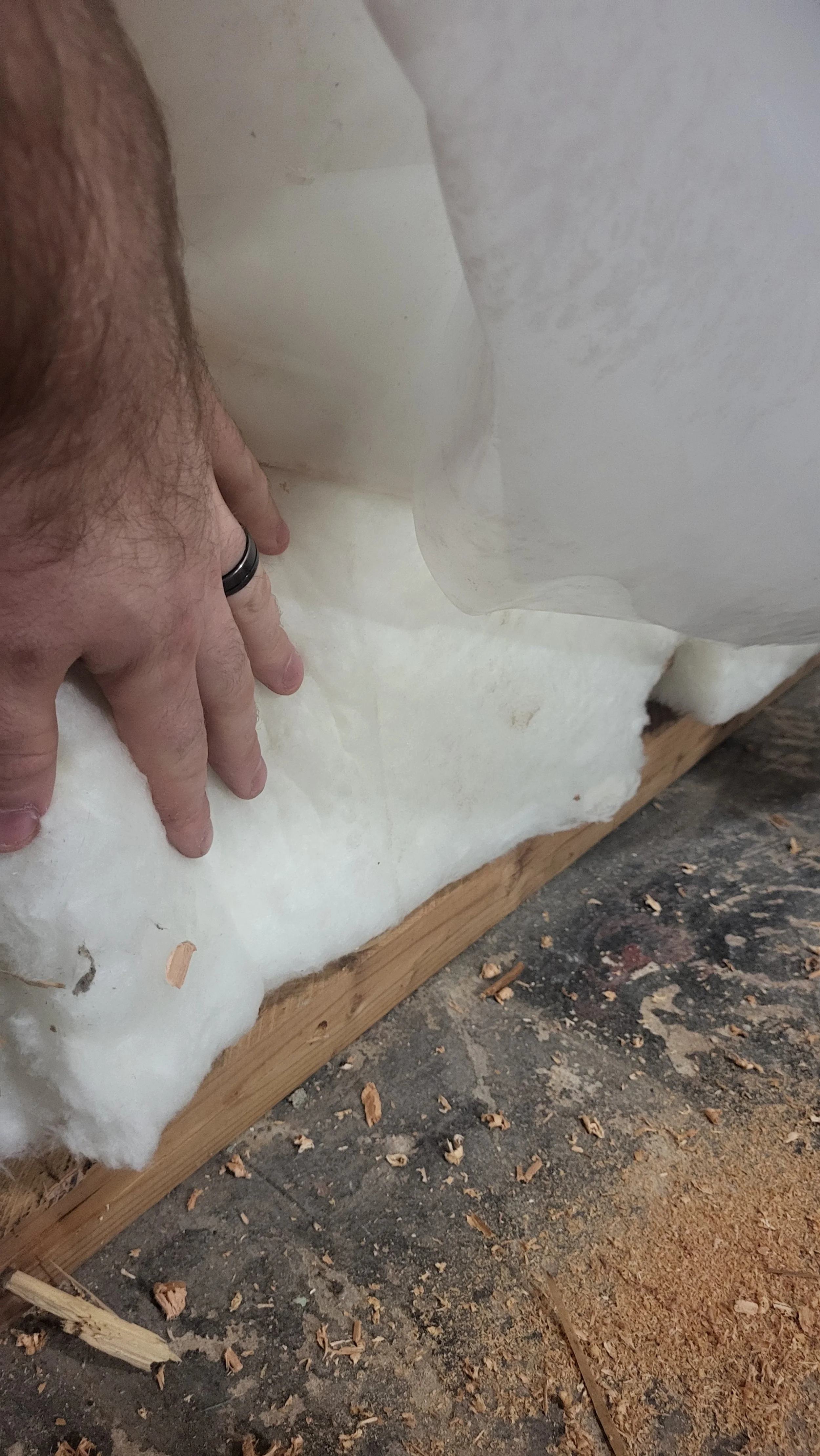 A person's hand with a wedding band smoothing out insulation material on a wooden surface, with sawdust and wood chips surrounding.