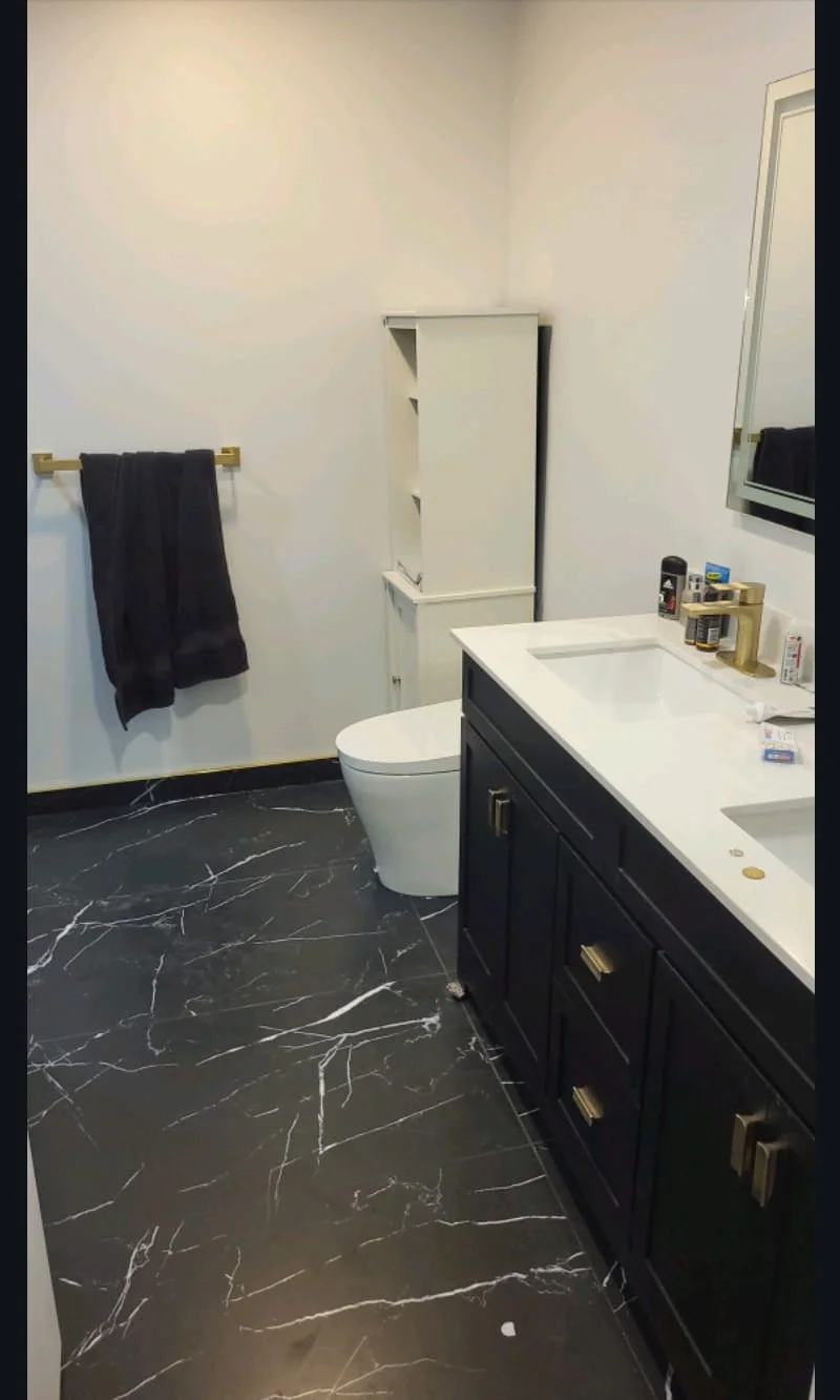 A modern bathroom with black marble floor, white walls, a black vanity with gold hardware, a white countertop, a black towel on a towel rack, a toilet, and a white cabinet. Bathroom contractor, tile installer, curb-less shower, 