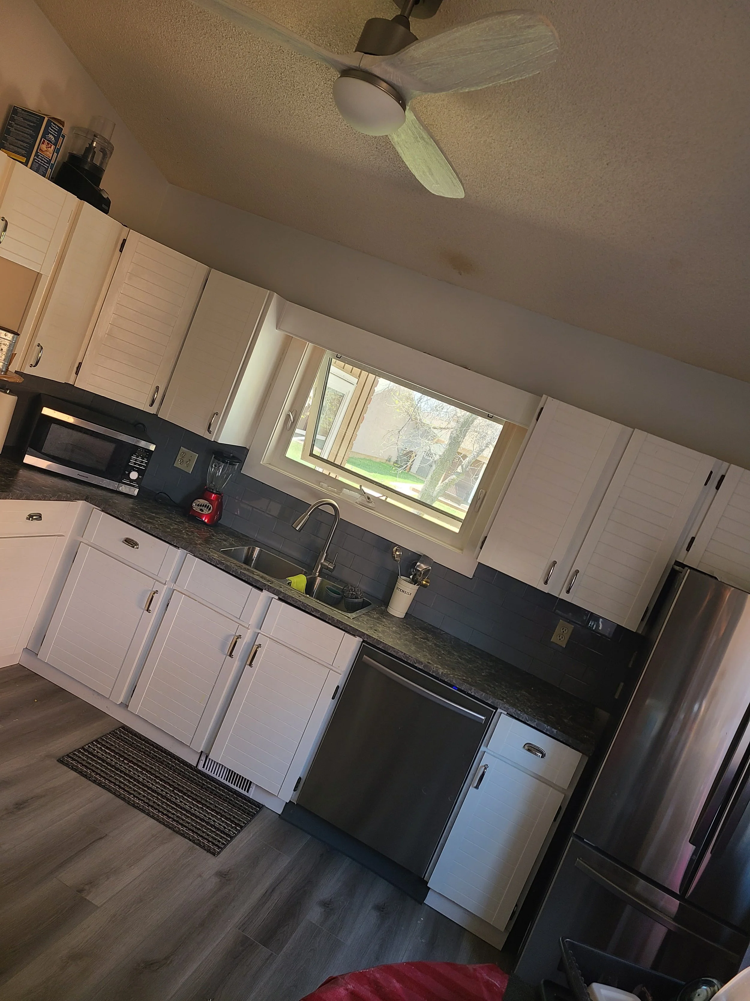 Kitchen with white cabinets, a black countertop, stainless steel appliances, a window above the sink, and a ceiling fan. kitchen contractor, cabinet installer, appliance installer, drywall contractor, contractor.