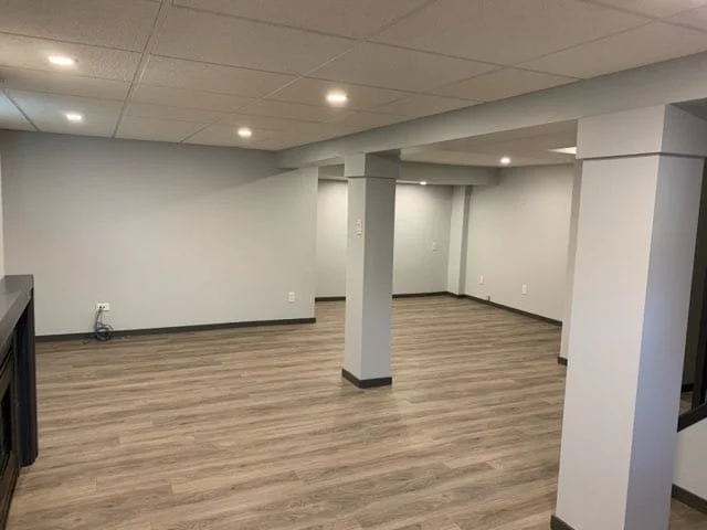 Empty basement or large room with light gray walls, wood laminate flooring, and recessed lighting in the ceiling, with some support columns and electrical outlets visible.  Basement Renovation, hardwood floors, construction, drywall contractor, paint