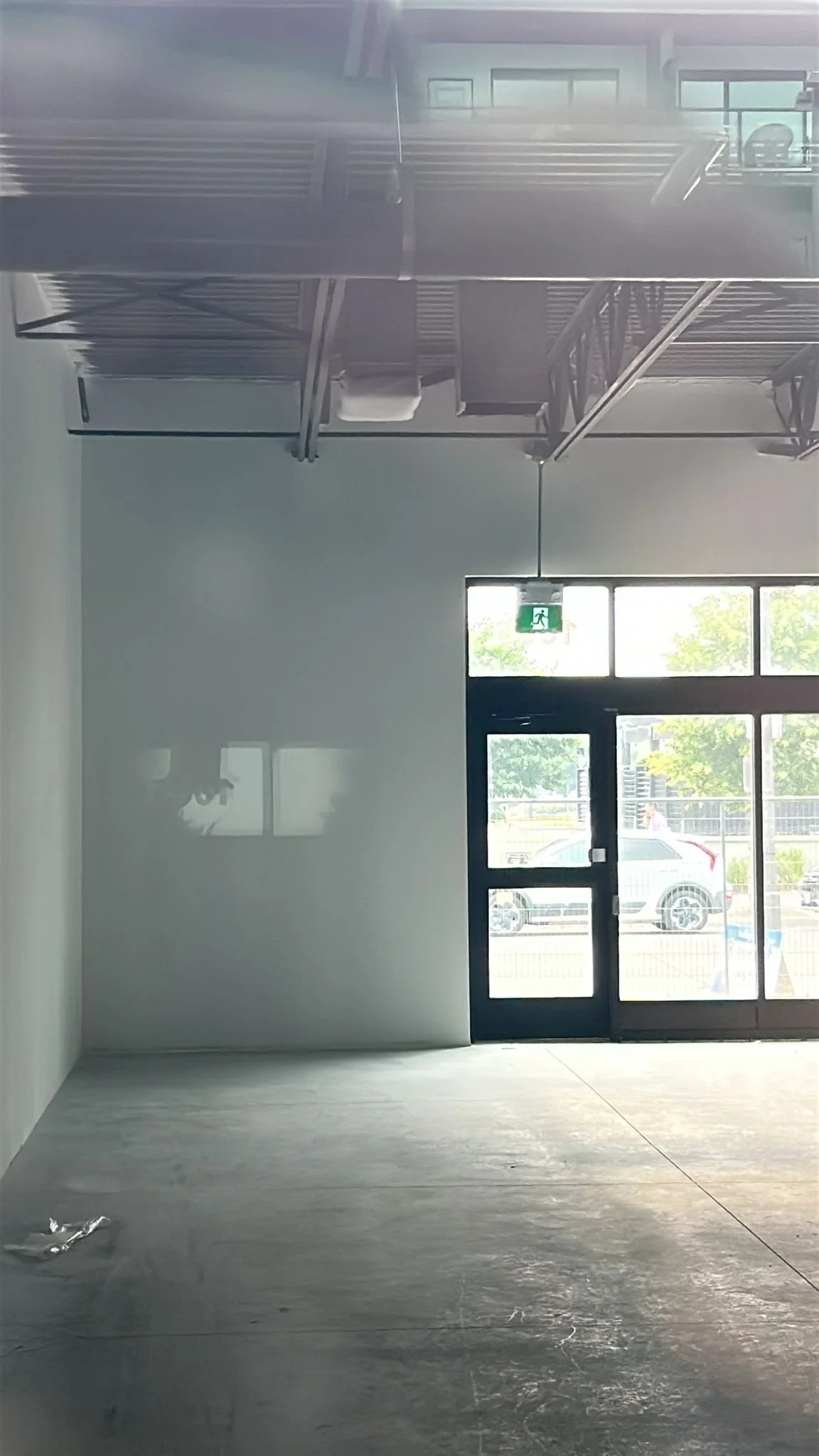 Empty store or office with glass doors and windows, exit sign above the door, some outdoor cars visible through the glass, and construction materials on the floor.