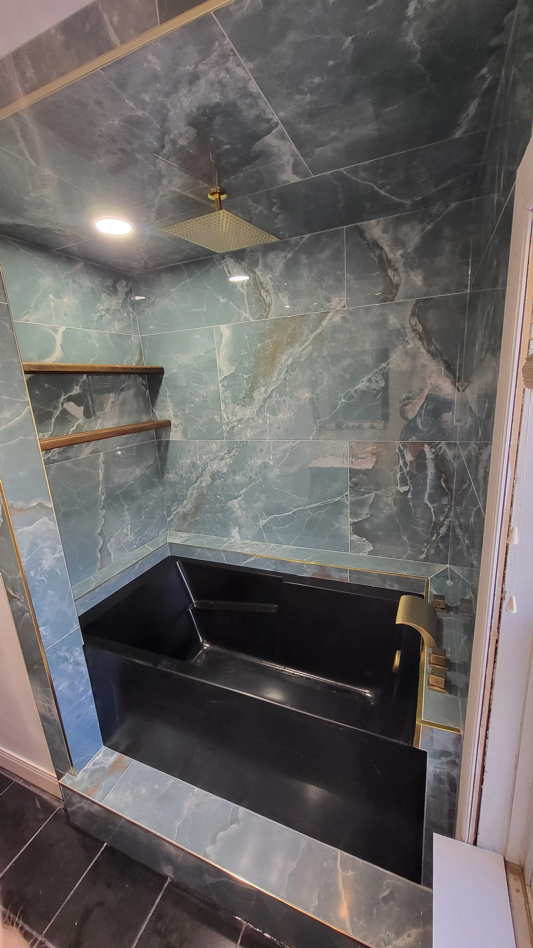 A modern bathroom shower with dark green marble tiles on the walls and ceiling, a black bathtub, wooden shelves, a square ceiling showerhead, and gold fixtures. Bathroom contractor, tile installer, curb-less shower, glass shower doors