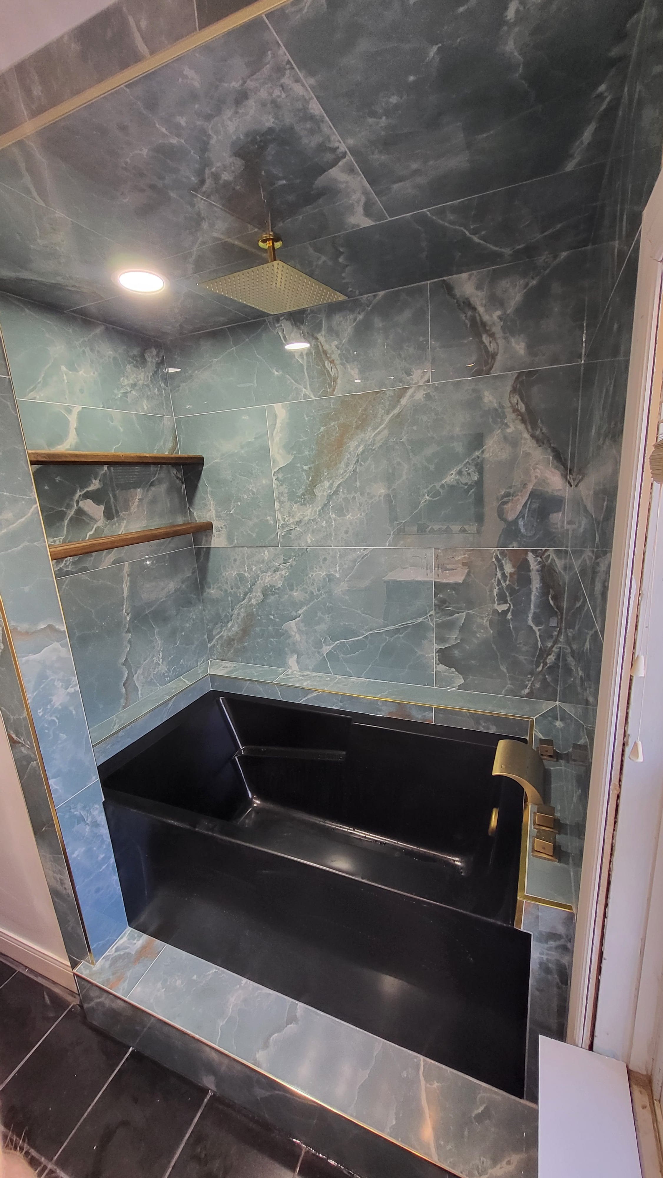 A modern bathroom shower area with dark green marble tiles on the walls and ceiling, a black bathtub, and gold fixtures. There are two wooden shelves on the wall and a rainfall showerhead mounted on the ceiling.