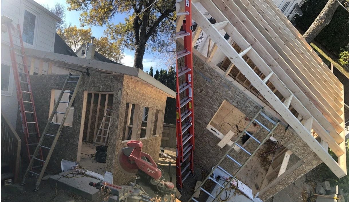 Side view of a house under construction with new roof framing and wall sheathing being added. Construction tools and ladders are visible around the site.
