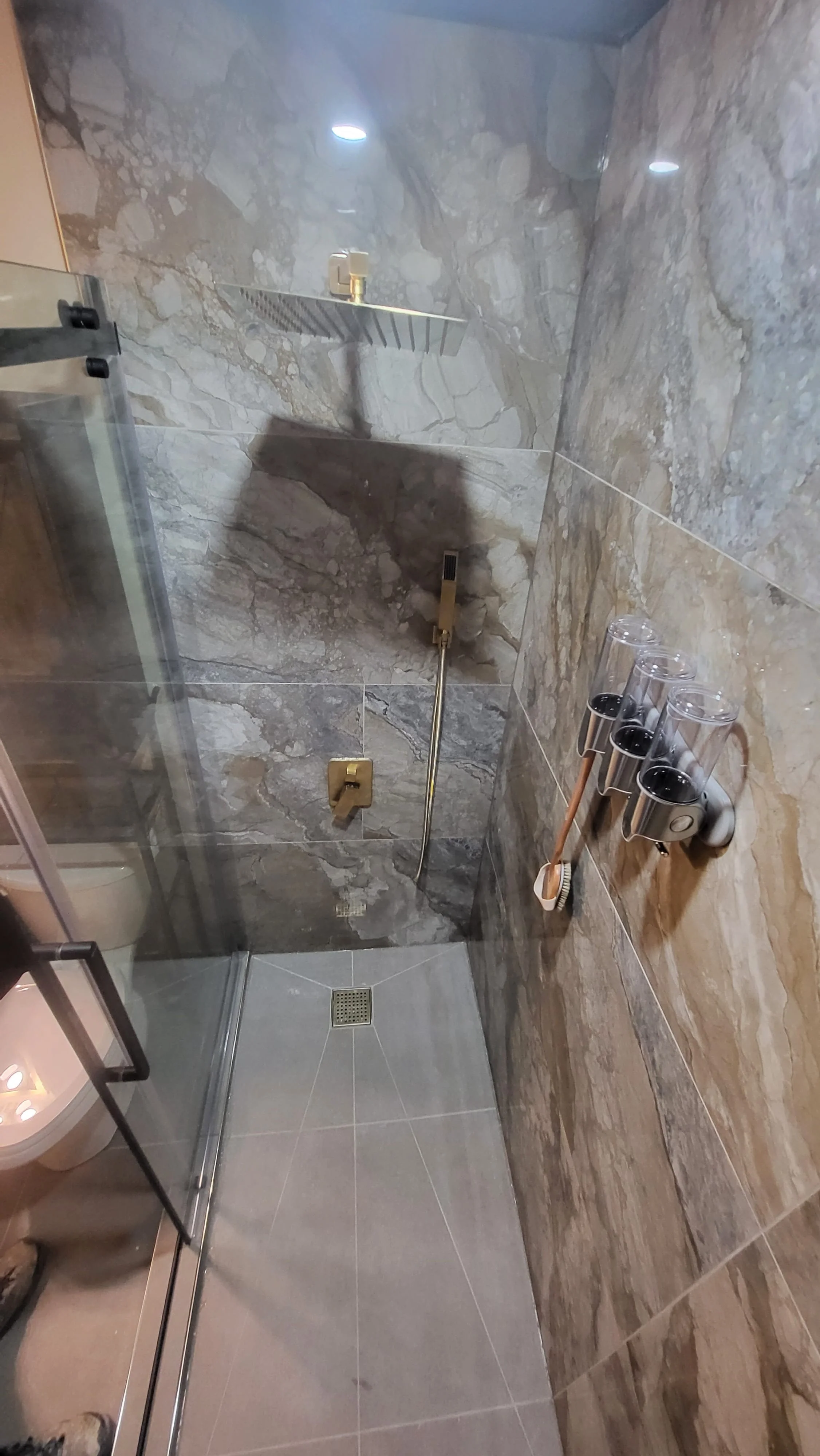 A modern walk-in shower with gray marble walls, a gold showerhead, a handheld shower, and a small square drain on the floor. Four glass cups with black bases are on a wall-mounted holder, alongside a mounted scrub brush. Bathroom contractor, tile ins