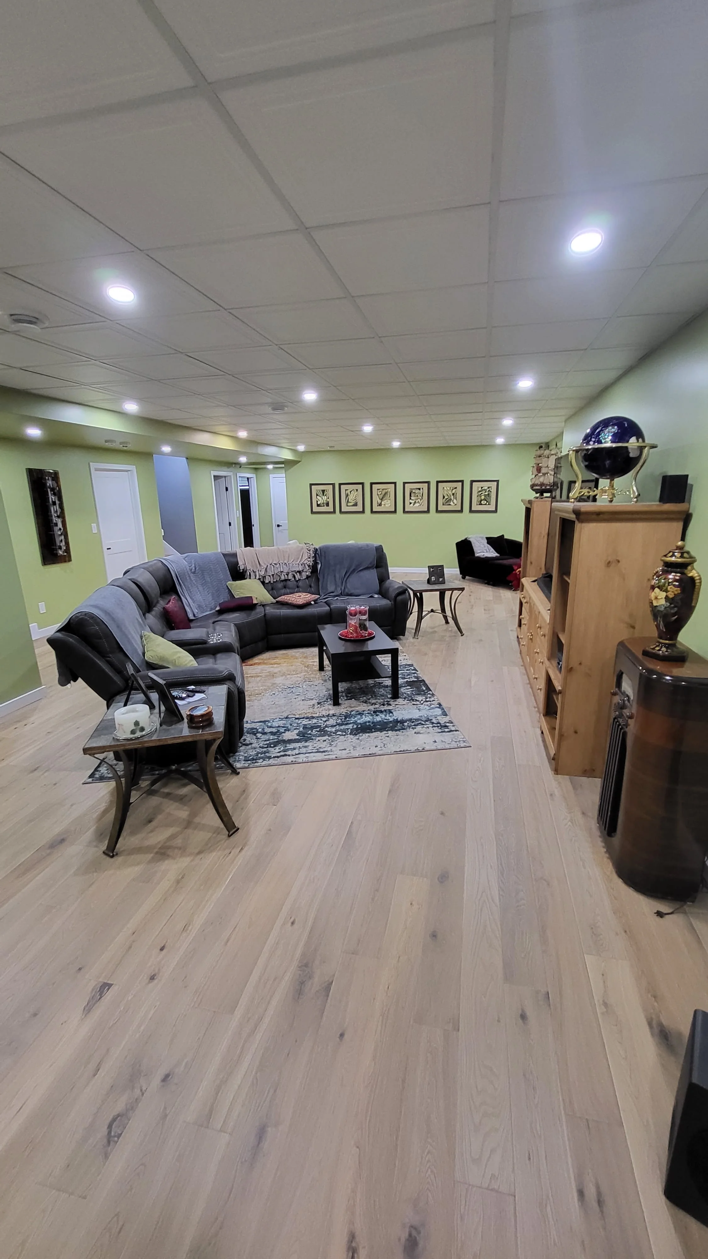 A living room with a black sectional sofa, small coffee table, and a wooden bookshelf. The room has green walls, framed artwork, and recessed ceiling lights. Basement remodel, hardwood floors.