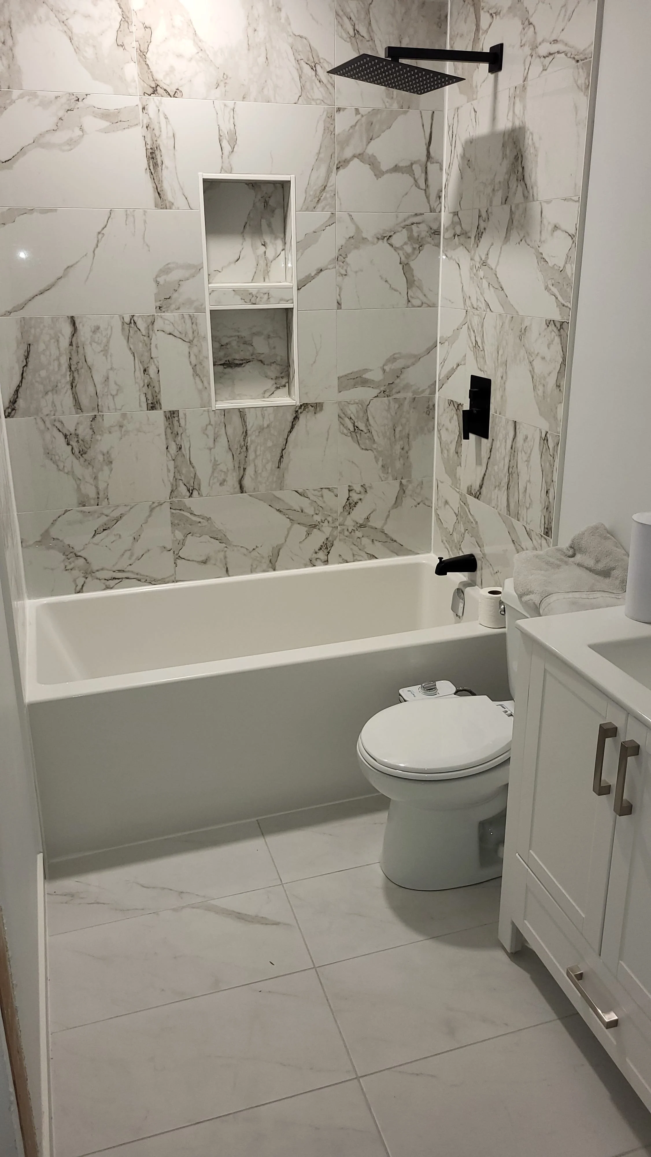 Bathroom with marble tiles, a bathtub, a toilet, a white vanity, folded towels, and a shower with a black rain showerhead and matching fixtures. Bathroom contractor, tile installer, curb-less shower, glass shower doors