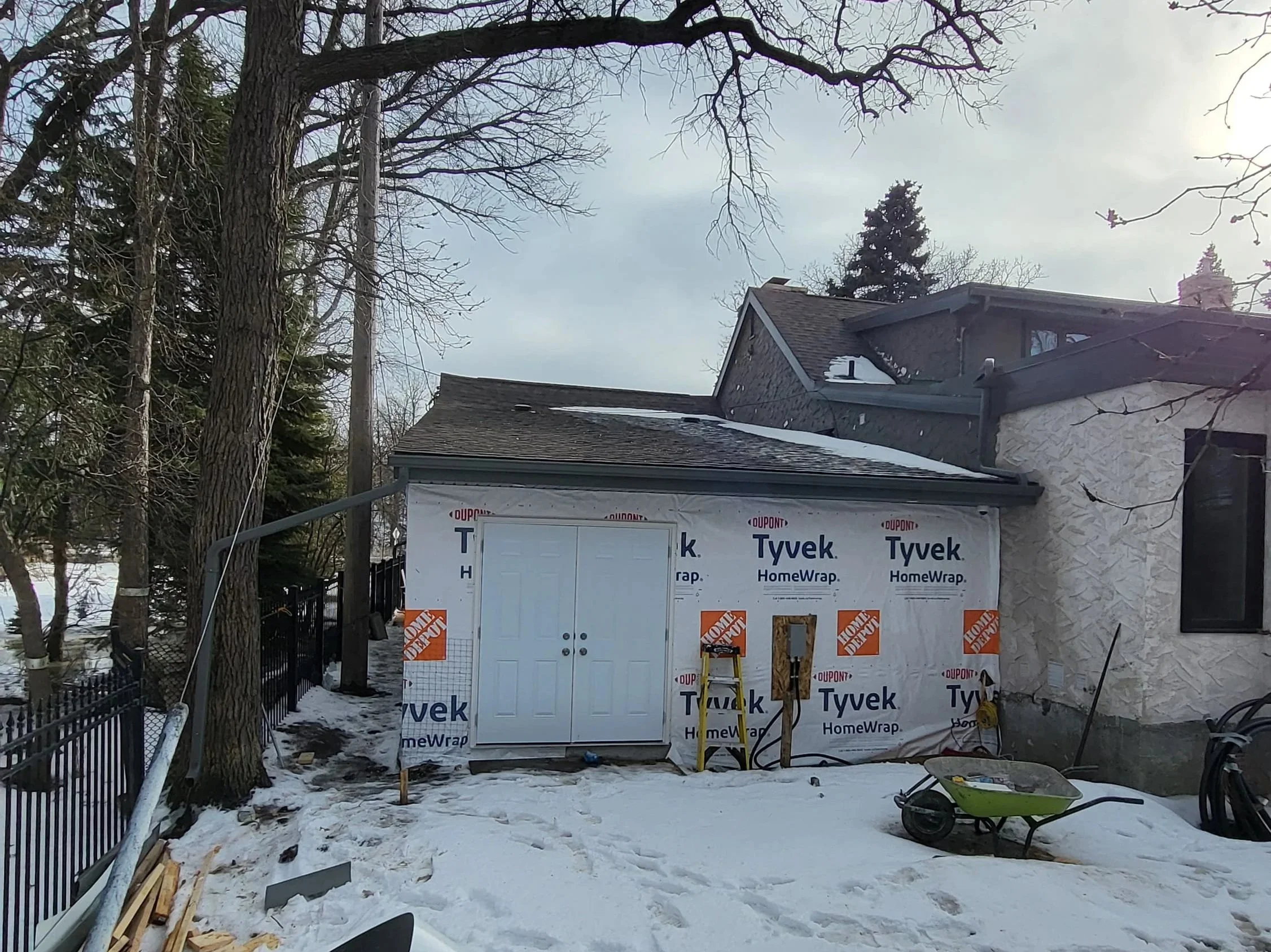 Side of a house under construction with white Tyvek HomeWrap, a small door, and a protective wheelbarrow in front, snow on the ground, and trees in the background. Home addition construction, garage extension.
