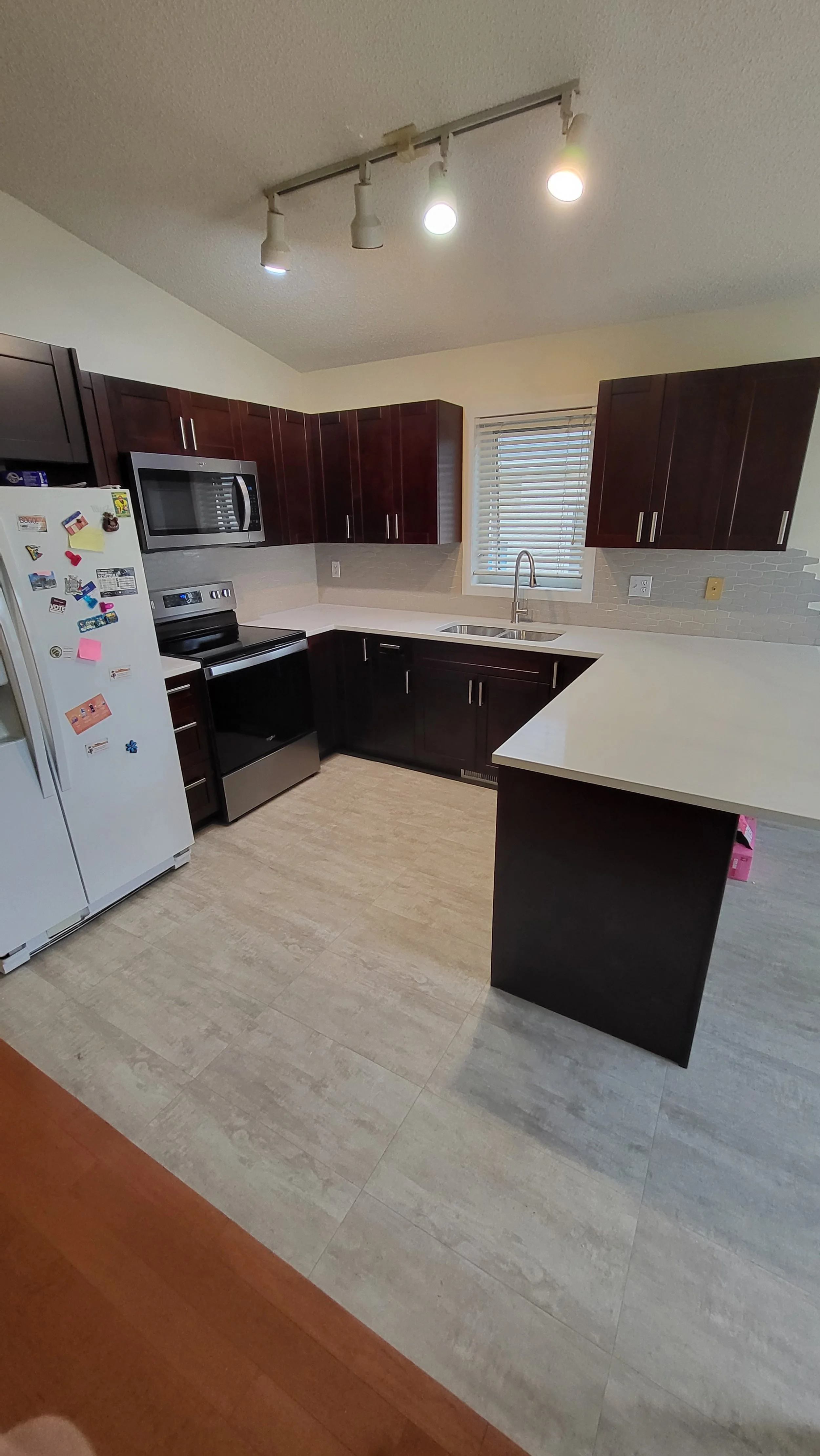 Modern kitchen with dark wood cabinets, white countertops, a stainless steel stove and microwave, white refrigerator with magnets, a small window with blinds, and track lighting on the ceiling. Kitchen remodel.