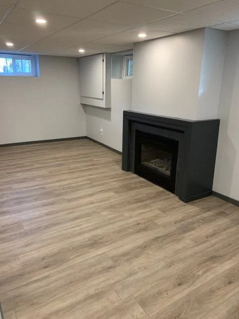 Empty room with light wood flooring, white walls, recessed ceiling lights, a black-framed fireplace, and small windows.  Basement Renovation, hardwood floors, construction, drywall contractor, painter, framer