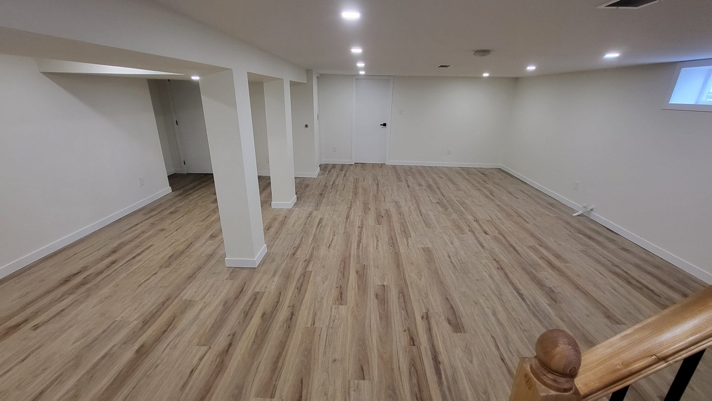 Empty finished basement with light wood flooring, white walls, recessed lighting, a small window, and a staircase with a wooden banister.  Basement Renovation, hardwood floors, construction, drywall contractor, painter, framer