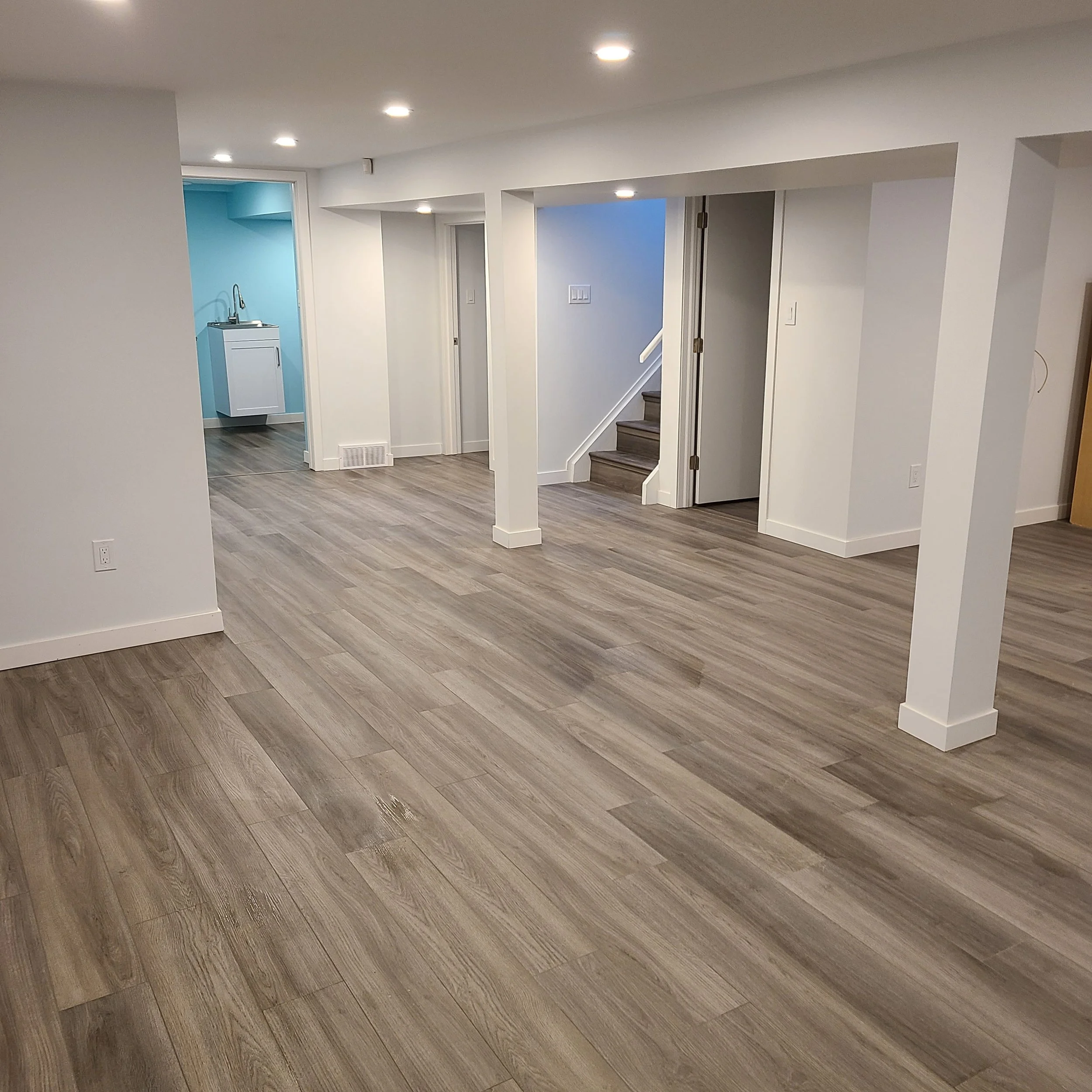 Empty room with wood flooring, white walls, recessed ceiling lights, a staircase, and a small room with a laundry station visible in the background.  Basement Renovation, hardwood floors, construction, drywall contractor, painter, framer