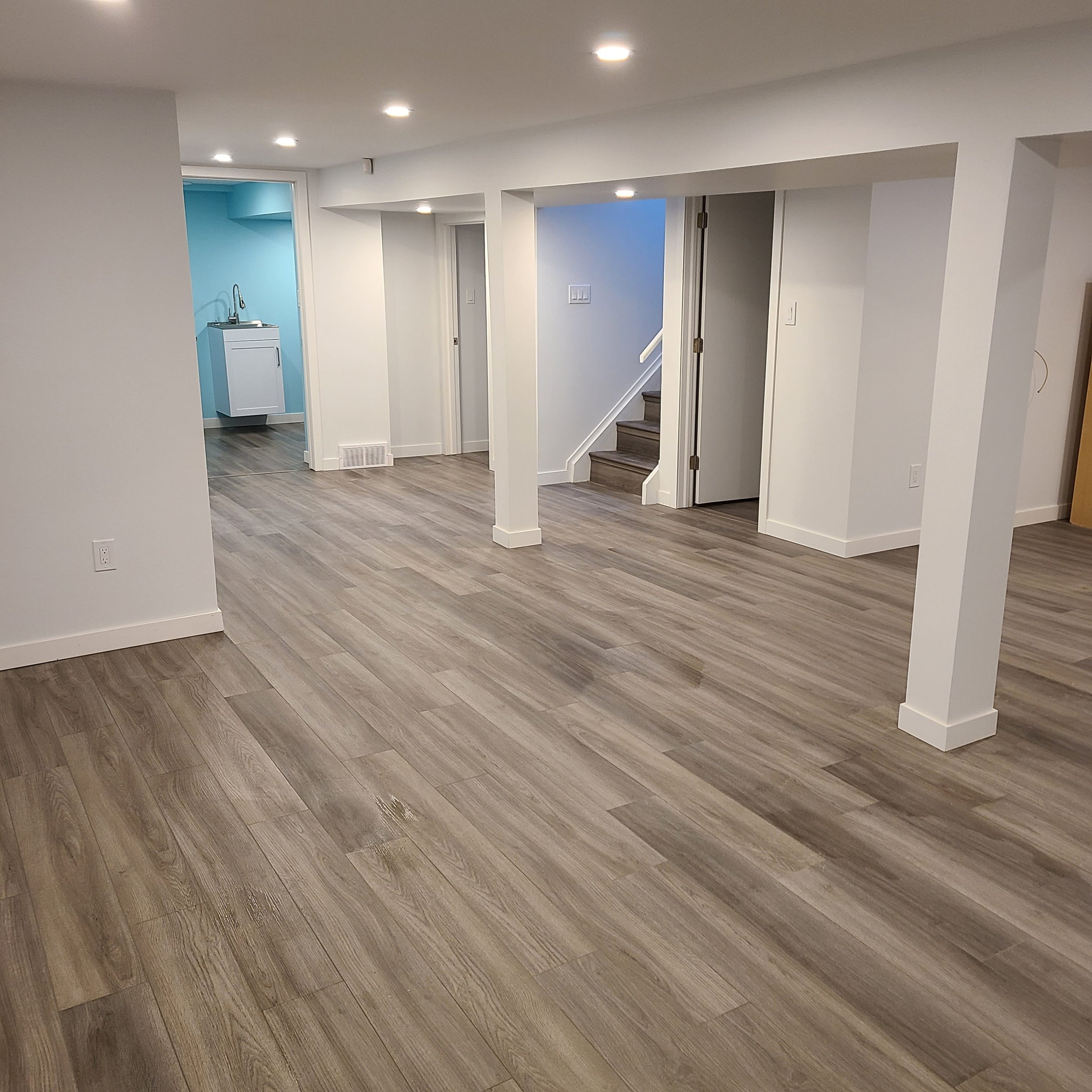 Empty room with wood-look flooring, white walls, and ceiling recessed lighting. Visible stairs and small laundry area in the background. basement remodel