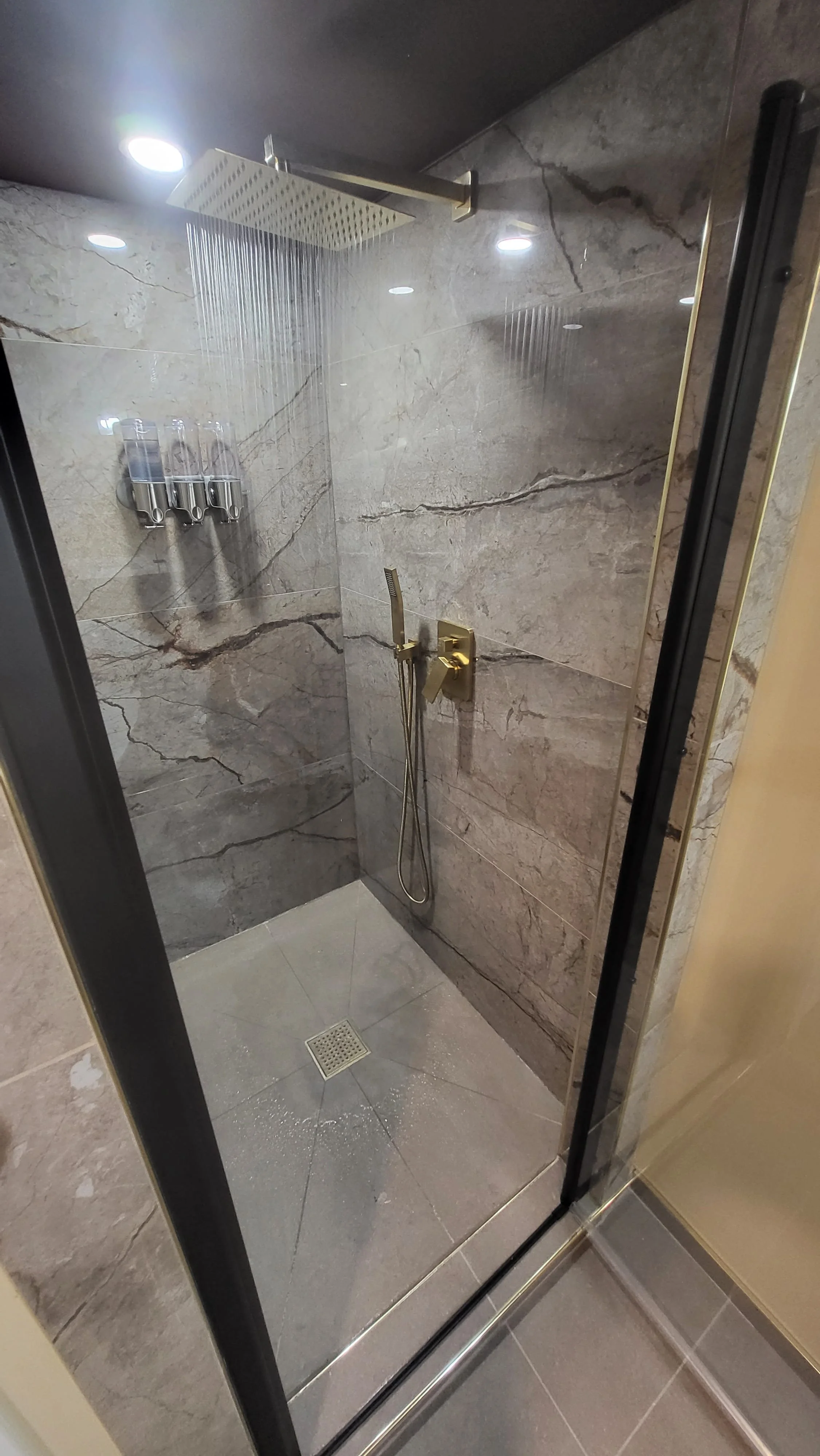 Shower with marble walls, gold fixtures, rainfall showerhead, handheld shower, and a square drain on the floor, viewed through a glass door. Bathroom contractor, tile installer, curb-less shower, glass shower doors