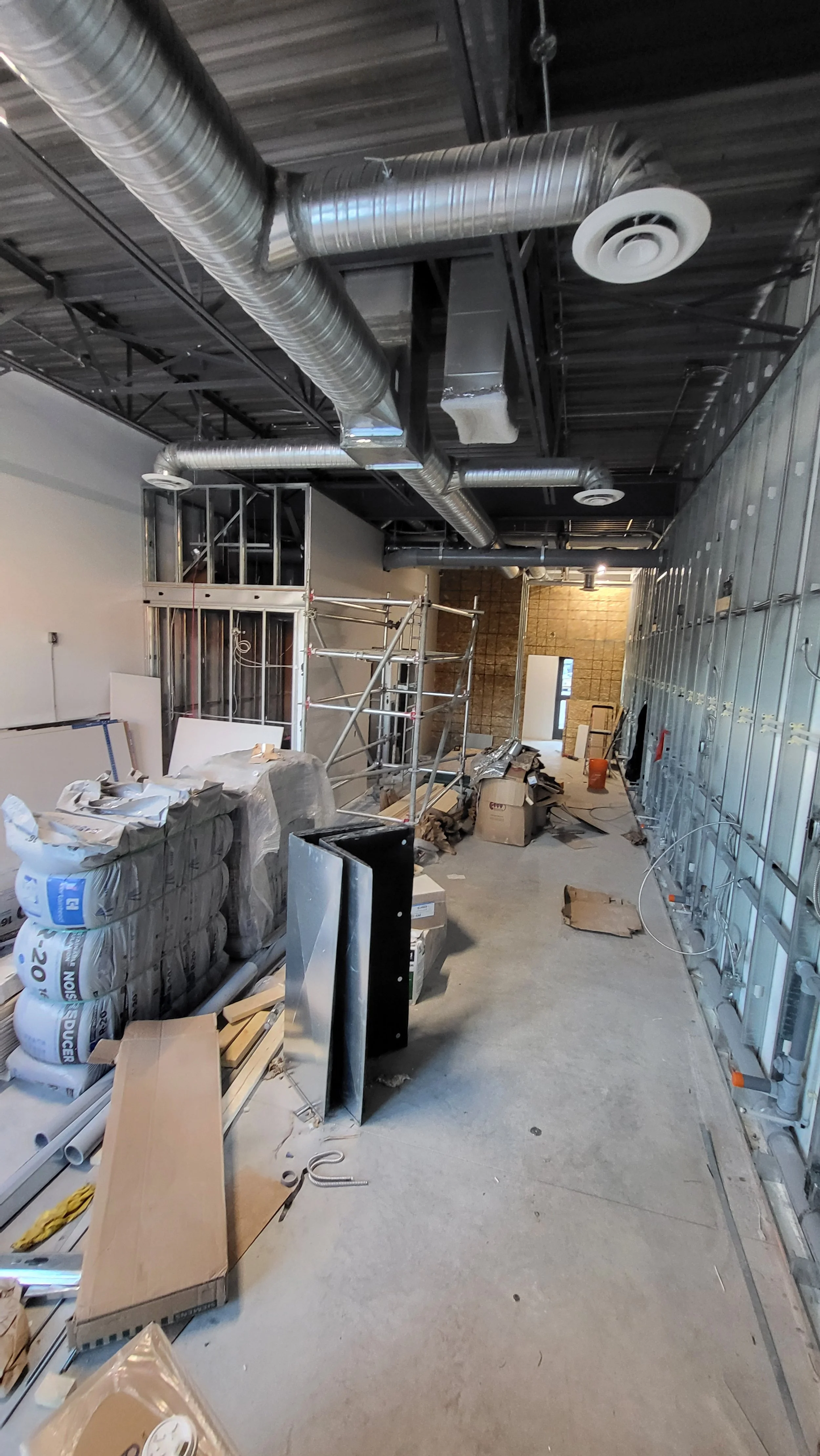 Interior of a building under construction with exposed ductwork, metal framing, scaffolding, and construction materials scattered on the floor.