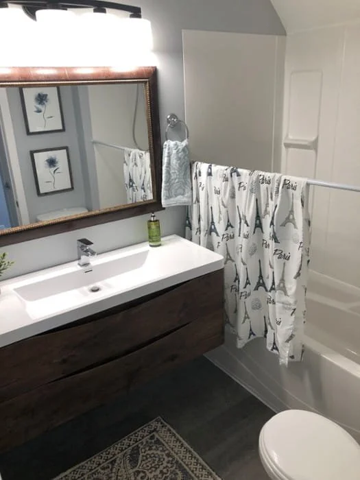 Bathroom with wooden vanity, white sink, mirror, framed art, towel, soap dispenser, shower curtain with Paris and Eiffel Tower designs, and bathtub. Bathroom contractor, tile installer, curb-less shower, glass shower doors