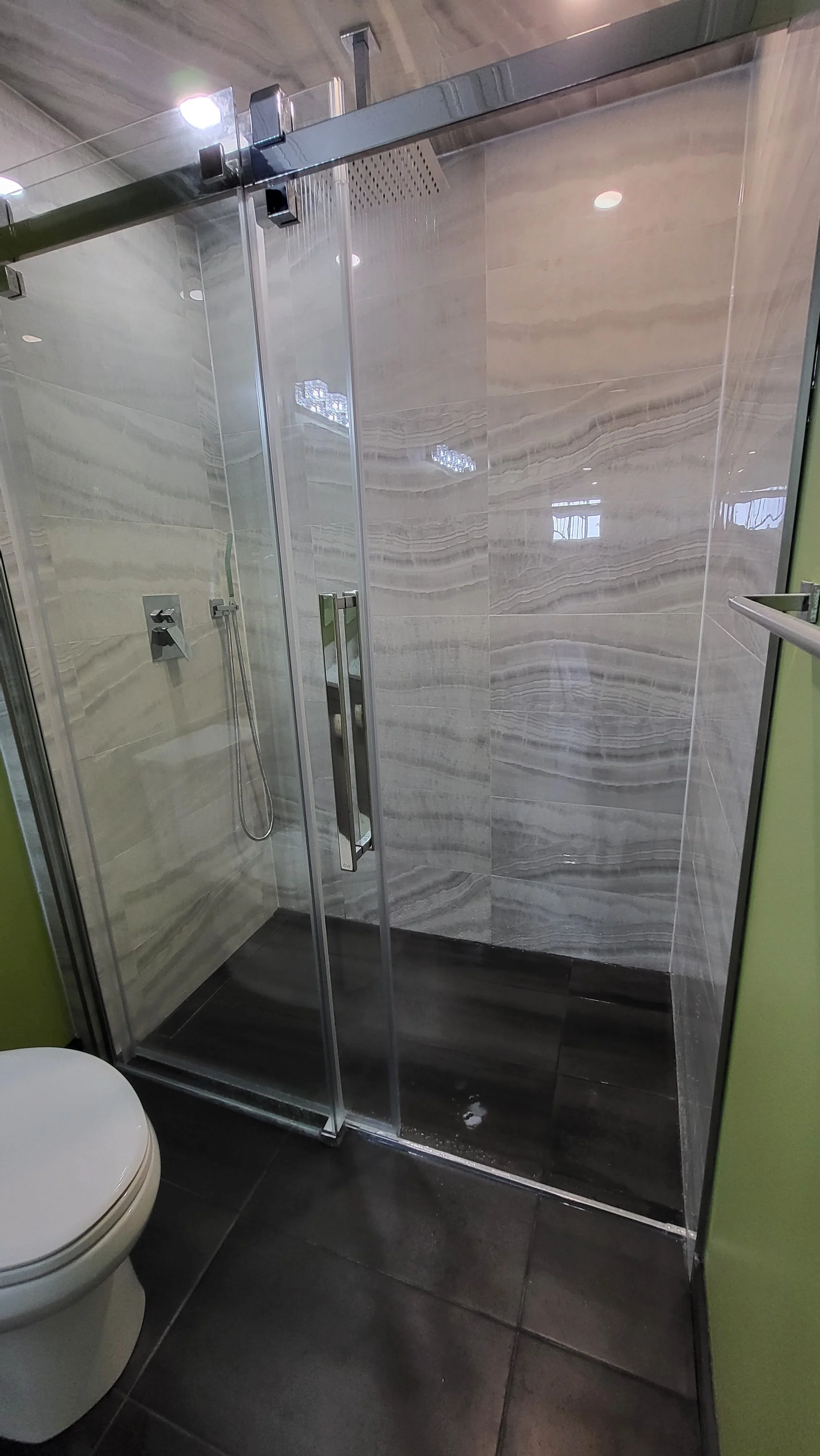A modern bathroom with a glass-enclosed shower area featuring marble-patterned tiles, a handheld showerhead, and a ceiling-mounted shower system, with a toilet visible in the foreground and a lime green wall on the right. Bathroom contractor, tile 