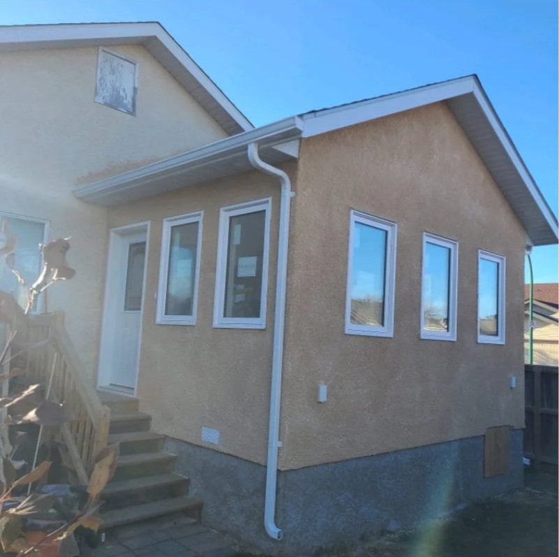 Exterior view of a house with beige stucco walls, a small staircase leading to a door, four windows on the side, and a white gutter system against a clear blue sky. Home addition construction.